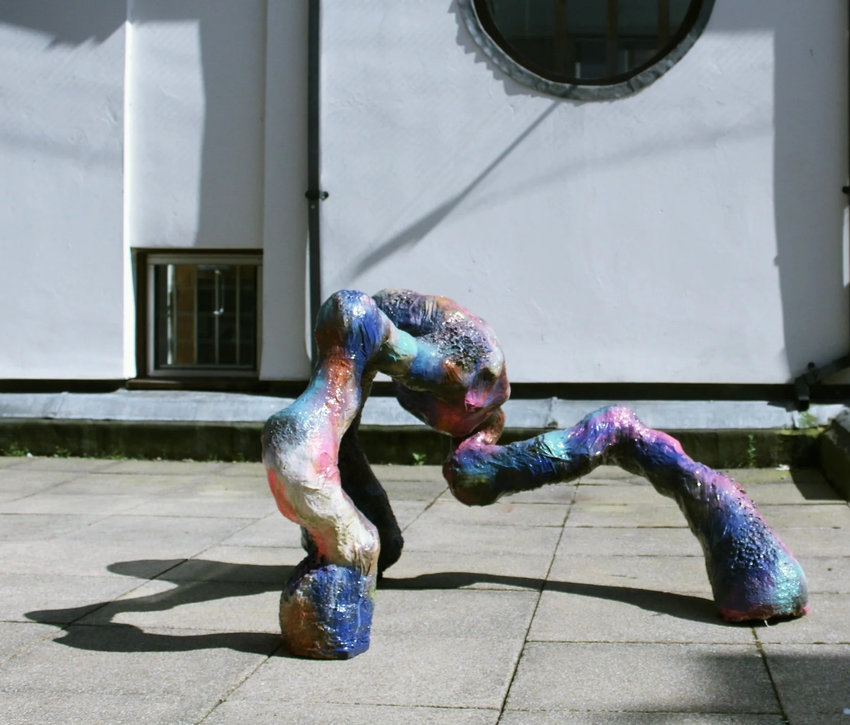 Colorful abstract sculpture on a sidewalk outside, casting a shadow.