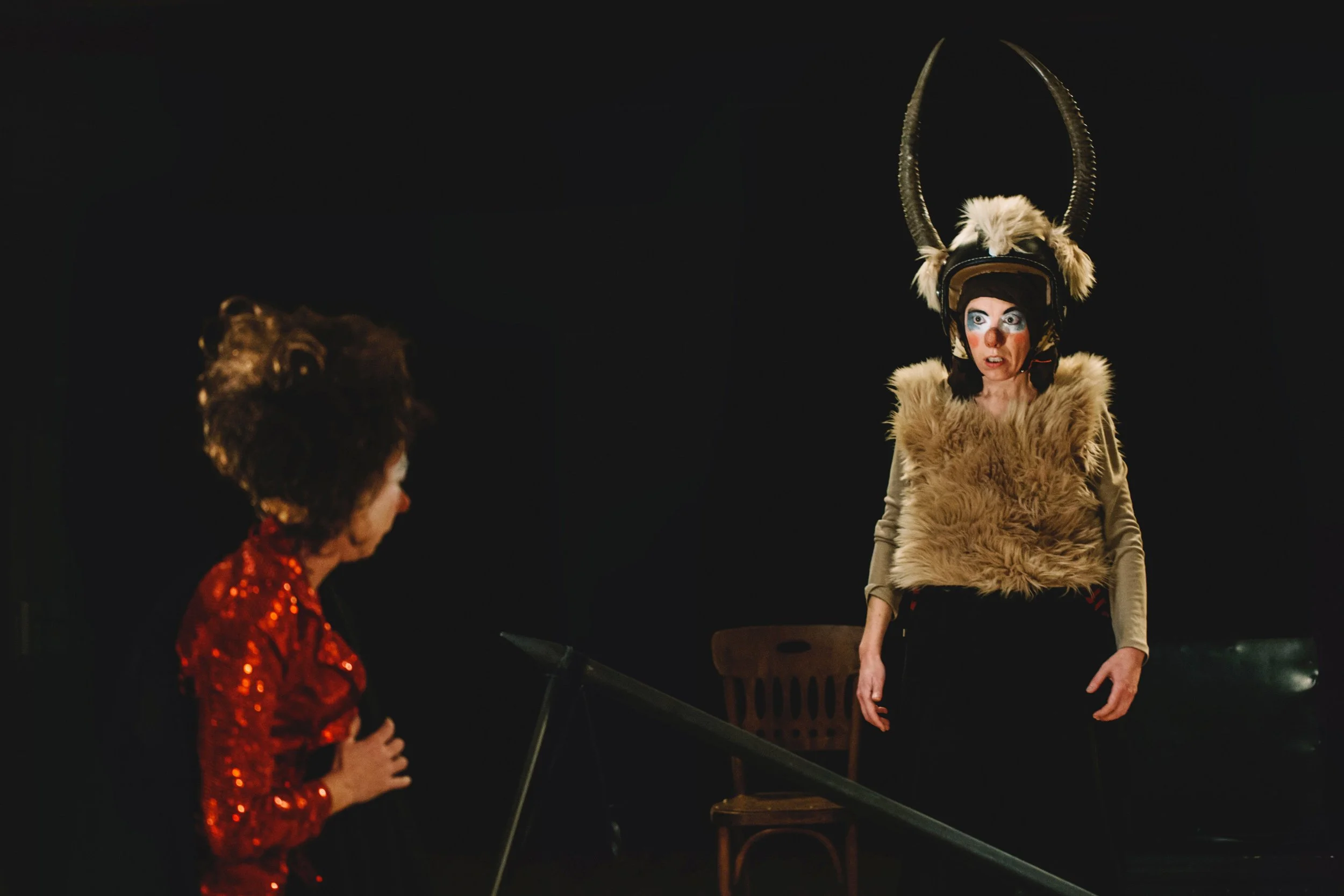 Une scène de théâtre avec deux femmes, une en costume de créature avec des cornes, porte un casque avec des cornes et un manteau de fourrure, l'autre en costume avec une robe rouge à sequins, dans une pièce sombre.
