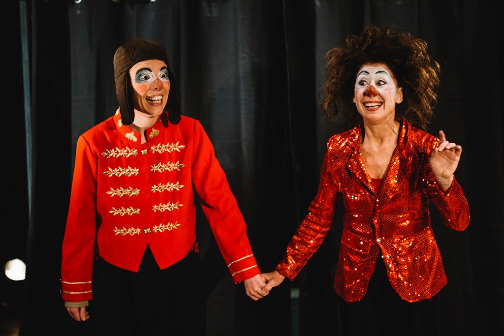 Deux femmes déguisées en clowns ou artistes avec costumes colorés, tenue rouge et maquillage facial, se tenant la main sur scène contre un fond noir.