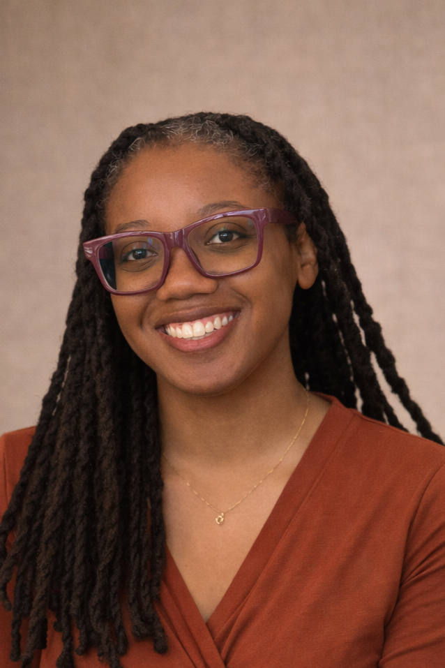 Sarie McGlone, A woman with dark skin, wearing purple glasses, smiling, with long dreadlocks, a gold necklace, and a brown top, standing against a neutral background.