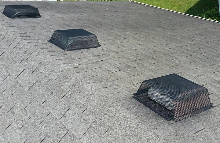 Three black vent covers on a gray asphalt shingle roof with a green yard in the background.