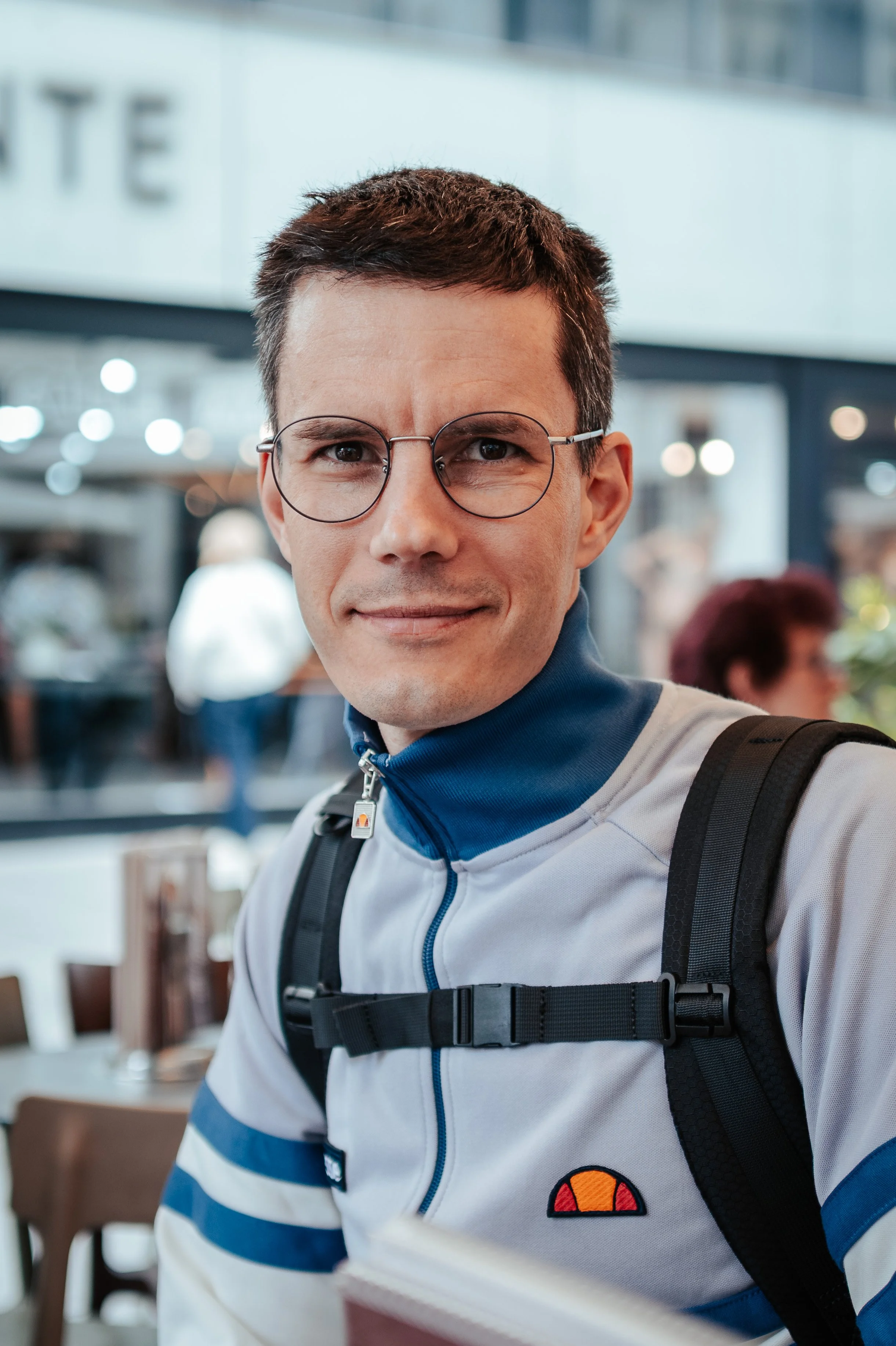 Junger Mann mit Brille, sportlicher Kleidung und Rucksack, sitzt in einem Café, lächelt in die Kamera.