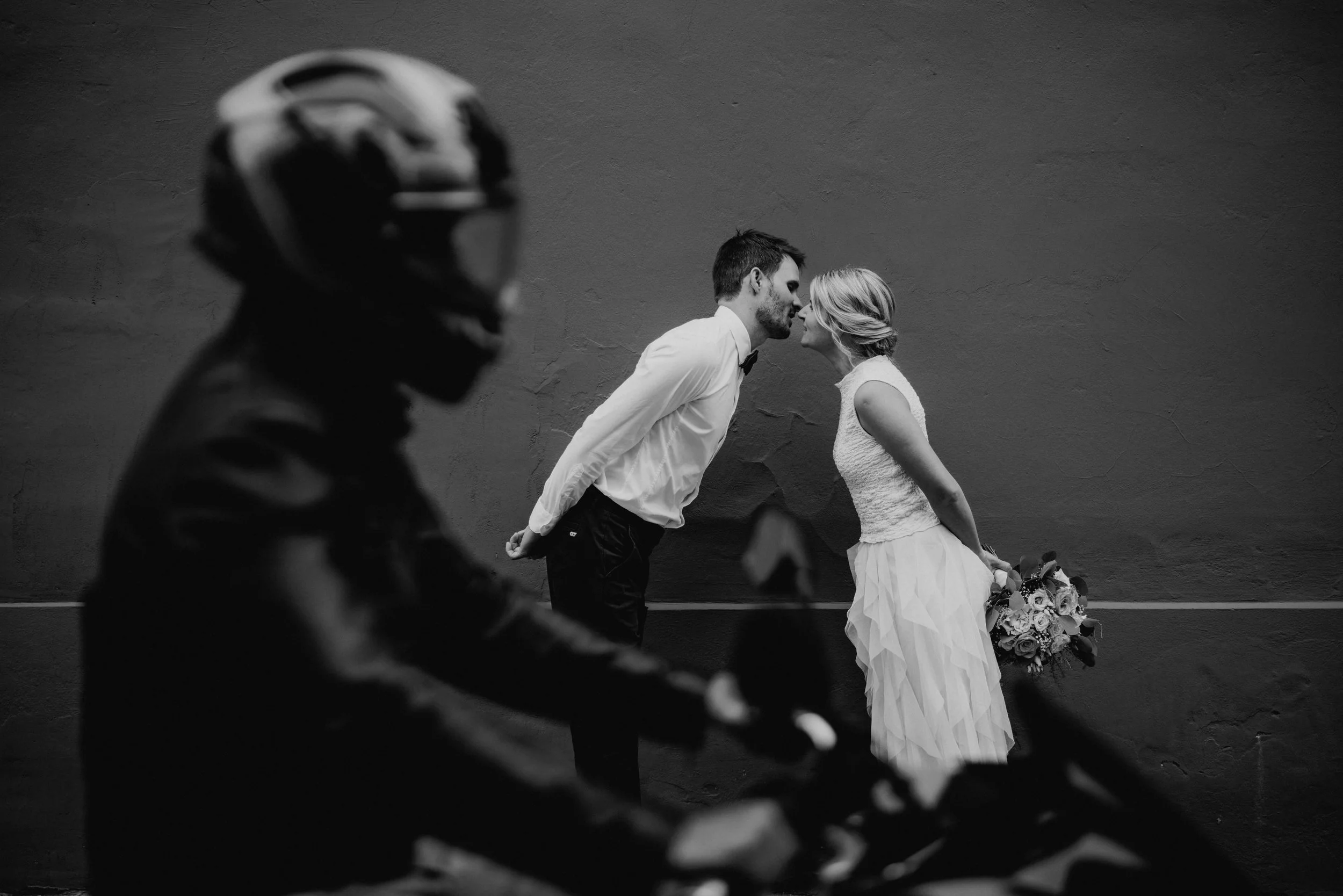 Ein Brautpaar steht sich vor einer dunklen Wand gegenüber, sie küsst sich. Im Vordergrund fährt eine Person mit Helm auf einem Motorrad vorbei.