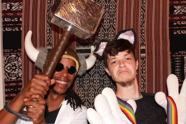 Two men dressed in costumes with a backdrop of patterned fabric. One man is holding a large hammer, wearing sunglasses and a Viking helmet with horns. The other man has a sad expression, wearing rainbow-colored suspenders and white gloves.