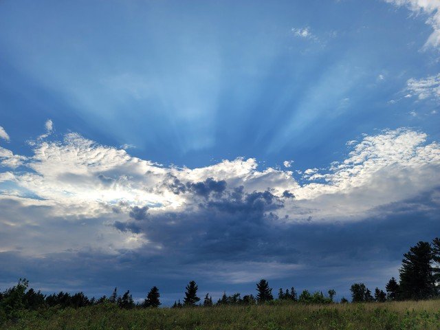 Sky with clouds and sunlight rays over a tree-lined field.