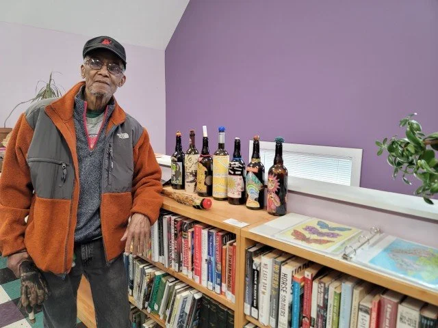 An elderly man stands next to a bookshelf and a table with several bottles of wine and a plant in a room with purple walls.