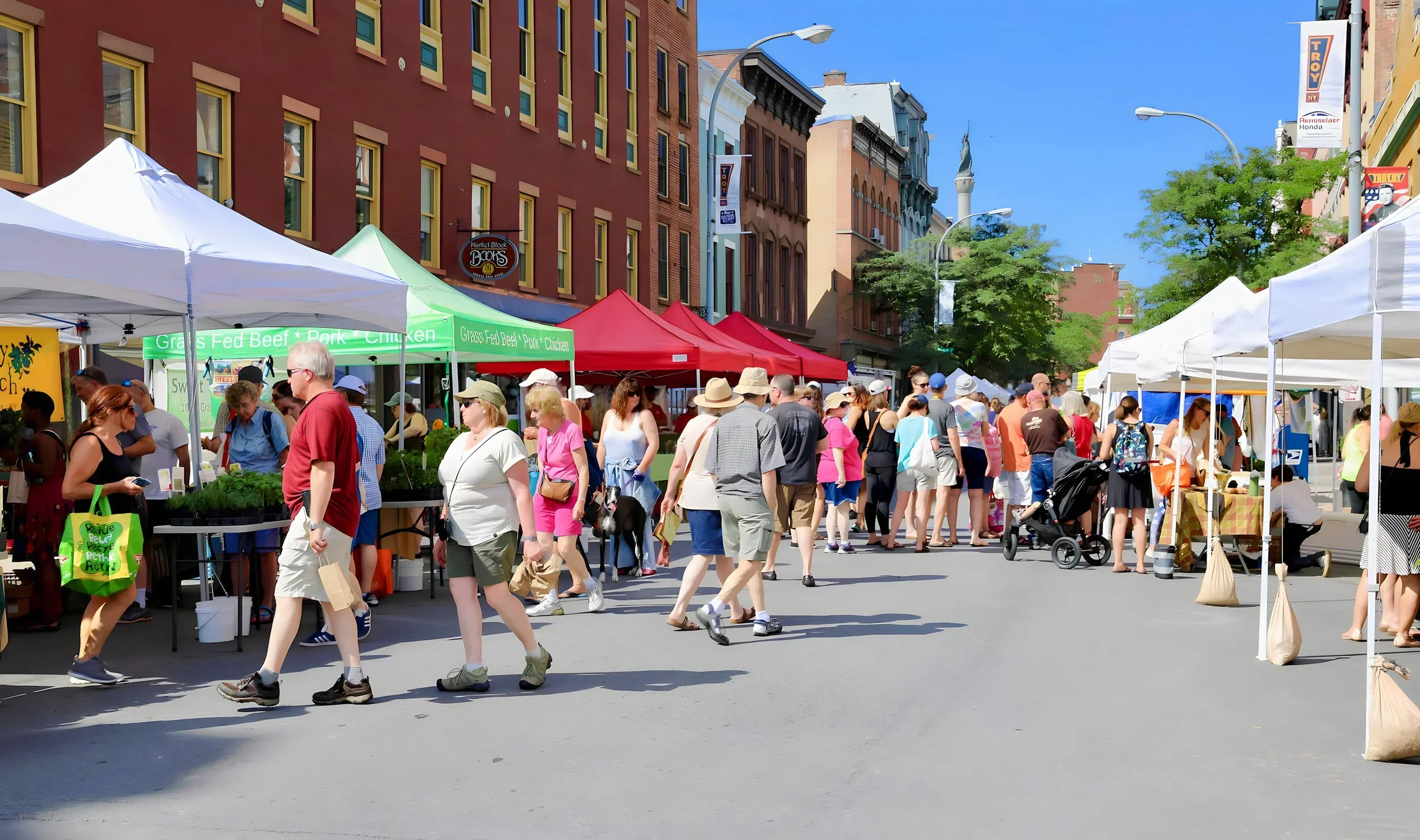 People shopping at outdoor market stalls on a city street with colorful buildings and blue sky.