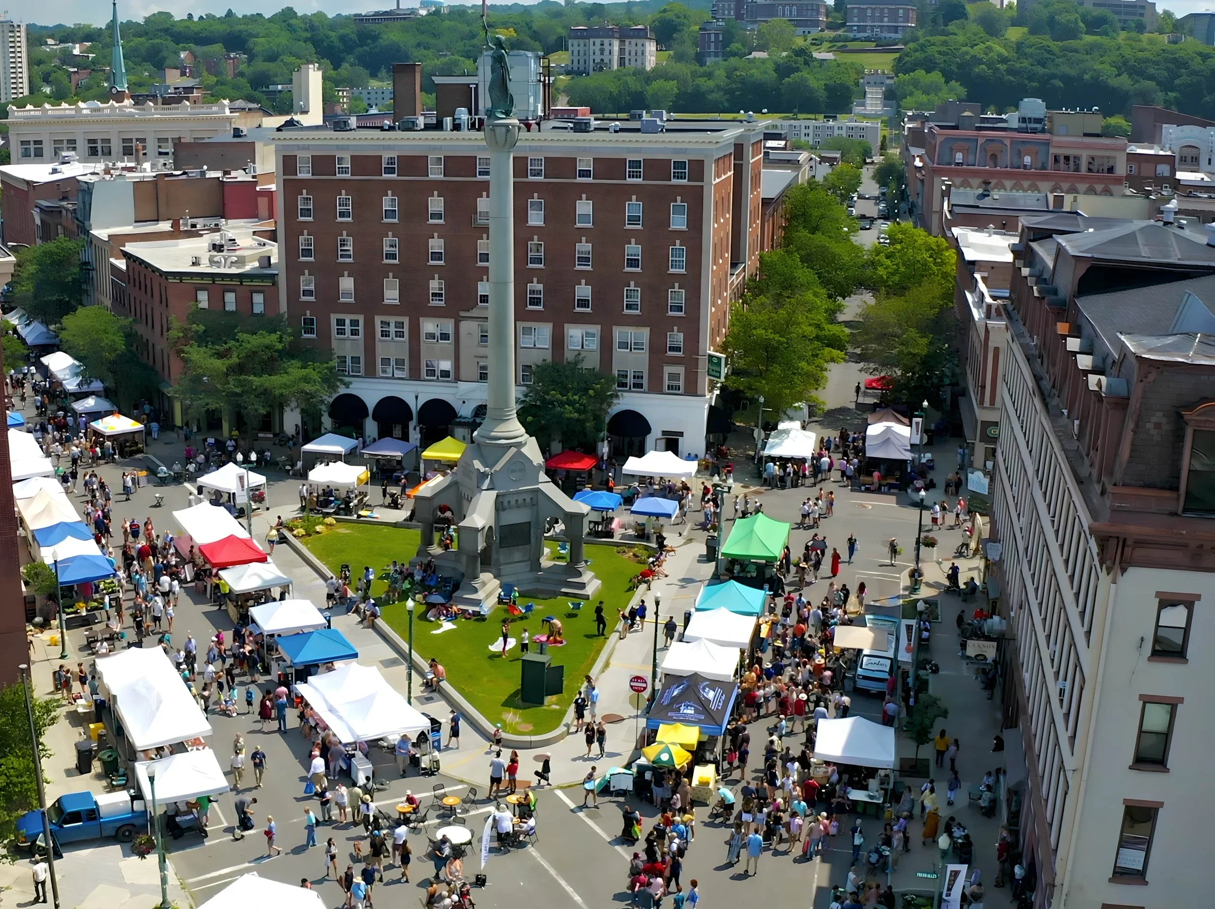 A bustling outdoor market in a city square with numerous vendors under colorful tents and a crowd of people walking and shopping, centered around a monument with a statue on top.