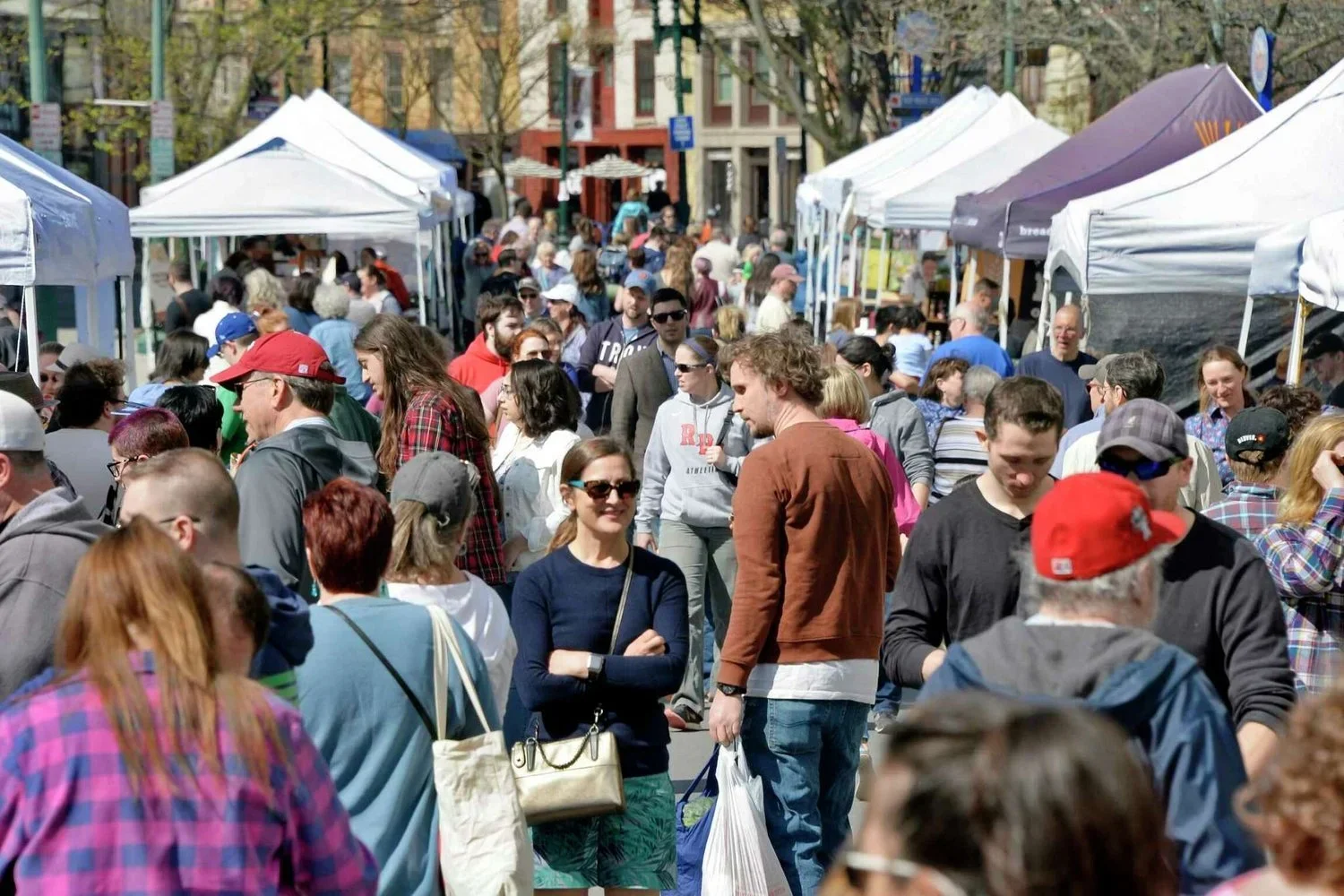 Crowd of people at an outdoor marketplace with white tents on a city street in daytime.