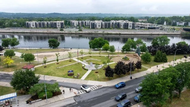 A park with walking paths, trees, and benches near a water body, with a street and cars in the foreground and modern apartment buildings across the water.
