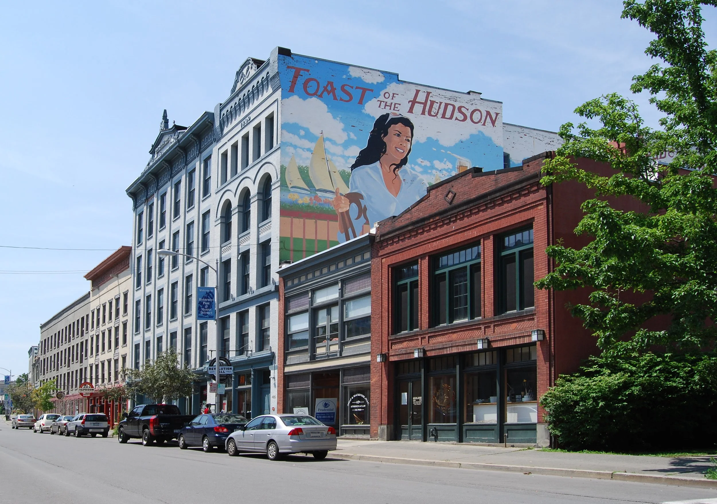 A city street with multi-story buildings, parked cars, a large colorful mural on a building depicting a woman and sailboats, and green trees.