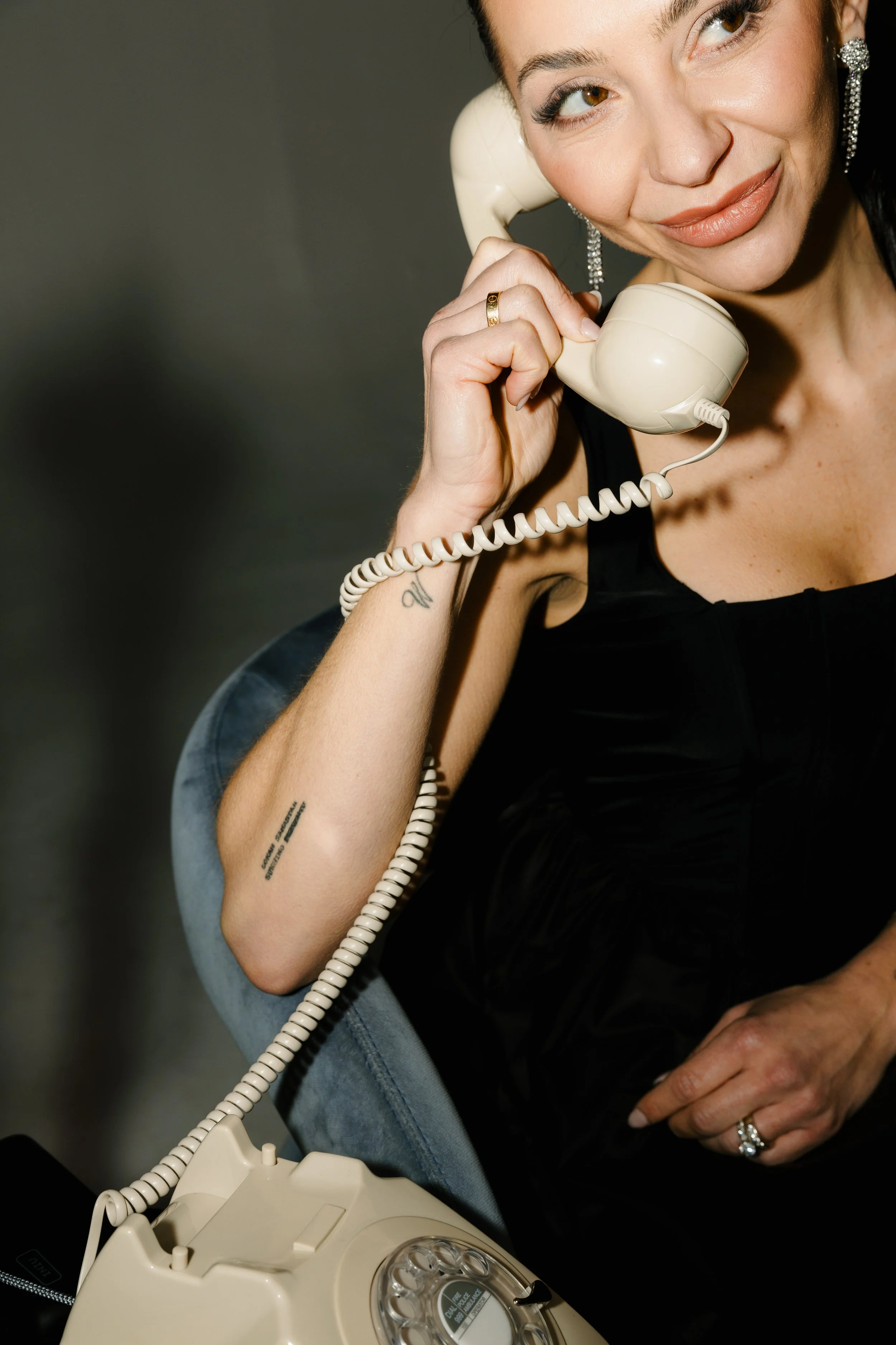 A woman with tattoos, wearing earrings and a black top, talking on a beige rotary phone.