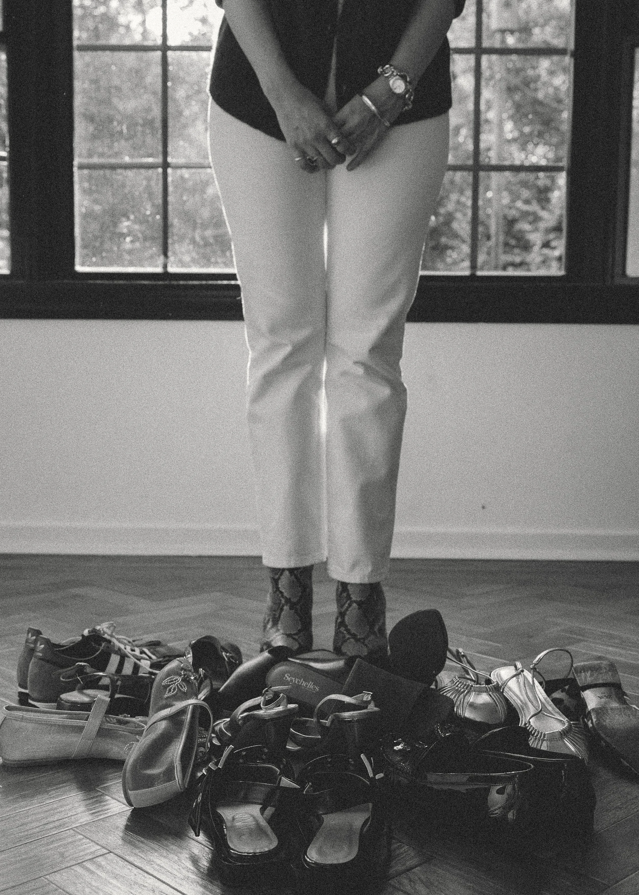 A person standing behind a collection of shoes on a wooden floor near a window with what appears to be a garden outside. The person is wearing white pants, patterned socks, and multiple bracelets.