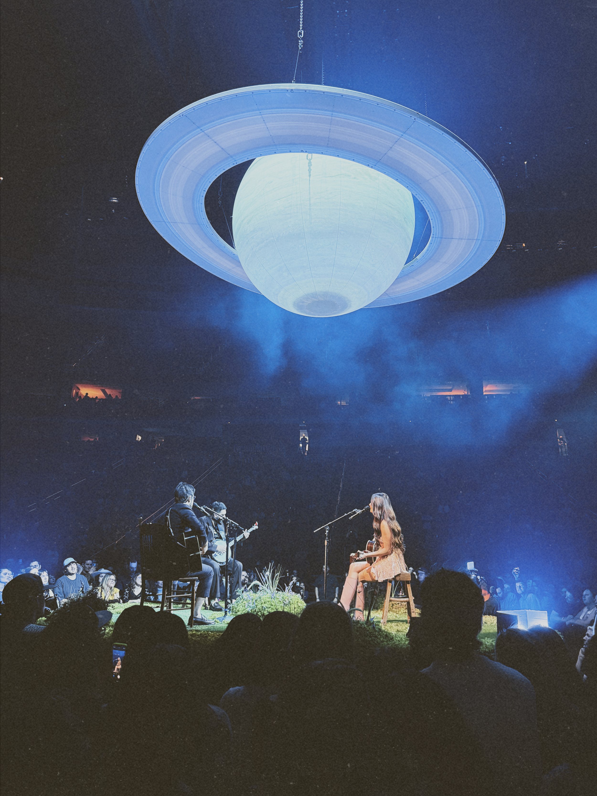 A musical performance on a stage with four musicians, three men and one woman, with a large Saturn-shaped structure hanging above them in a dark venue filled with an audience.