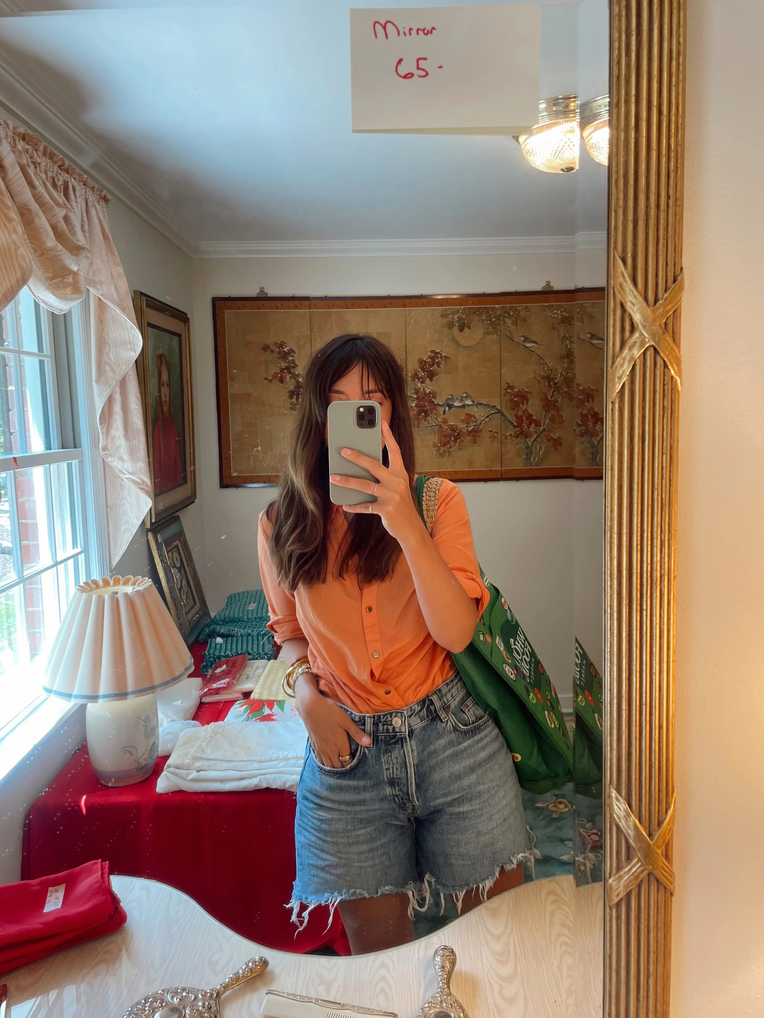 A woman taking a mirror selfie in a mirror with a gold frame. She is wearing an orange button-up shirt and denim shorts. She has a green tote bag over her shoulder. The room has a window with curtains, a dresser with a lamp, and vintage artwork on the walls. A note on the mirror reads "Mirror 65".