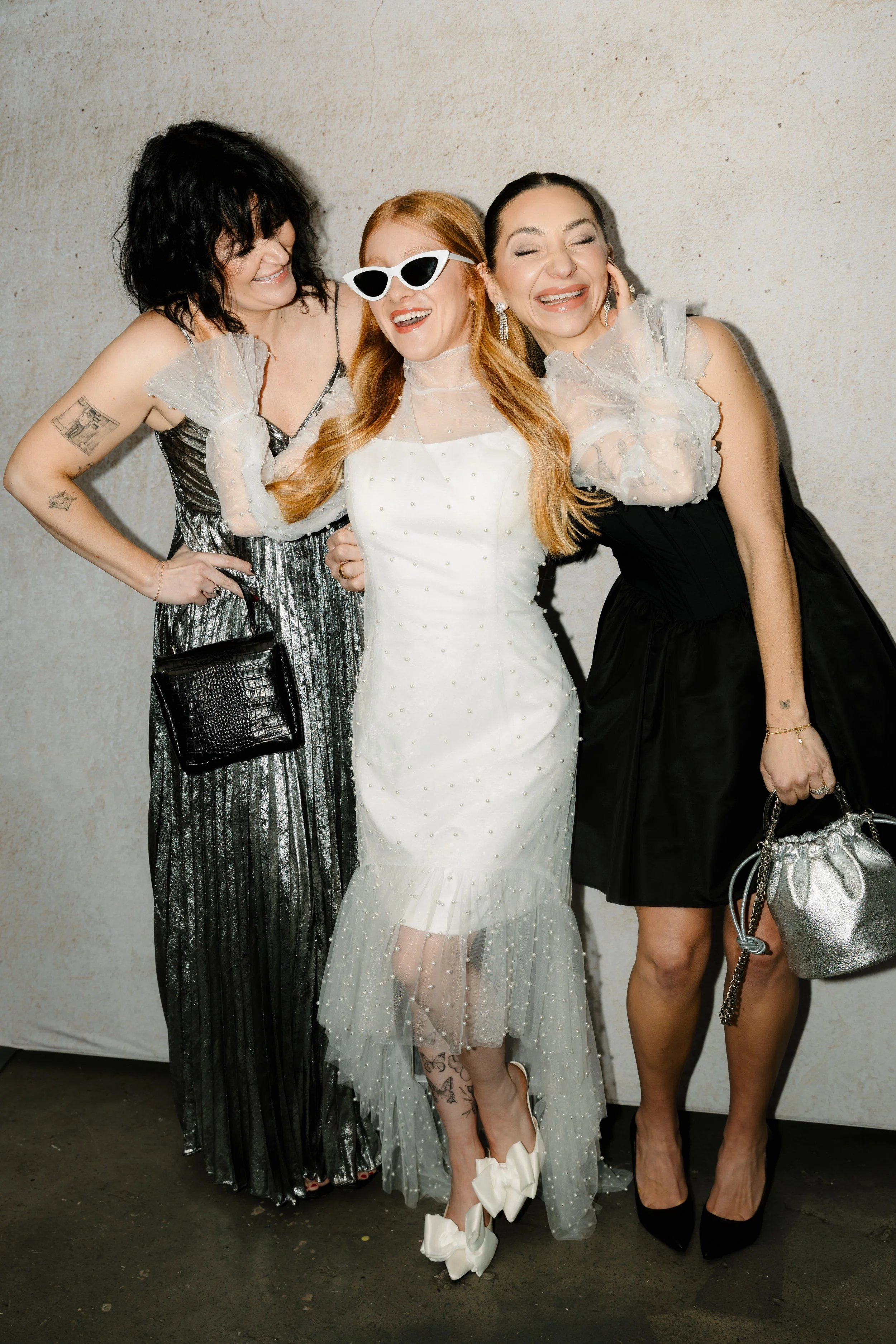 Three women dressed in elegant attire, smiling and celebrating together at an event, standing against a neutral background.