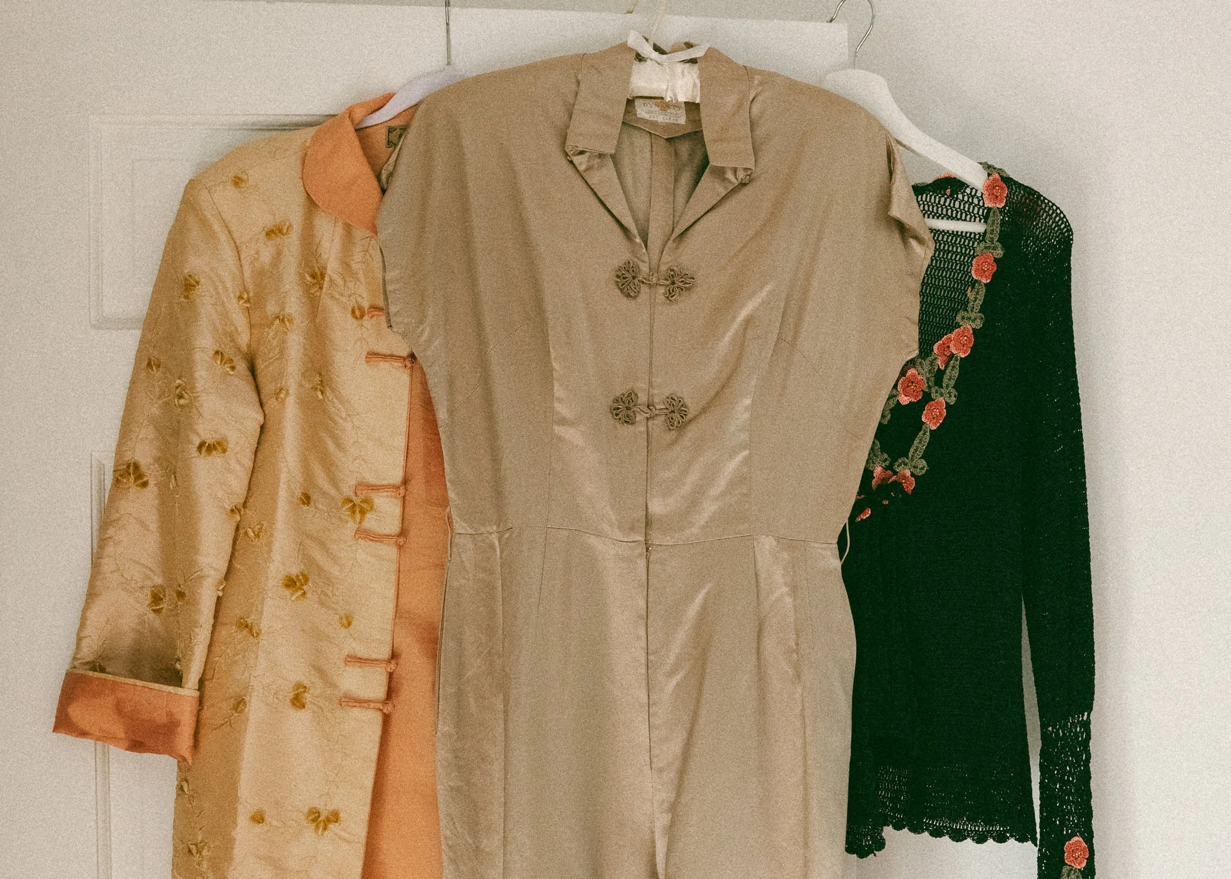 Three vintage dresses hanging on hooks against a plain wall. The first dress is peach with gold floral embroidery and satin trim. The second dress is cream-colored with a satin finish and decorative floral clasps. The third dress is black with open knit pattern and red floral embroidery along the sleeve and bottom.