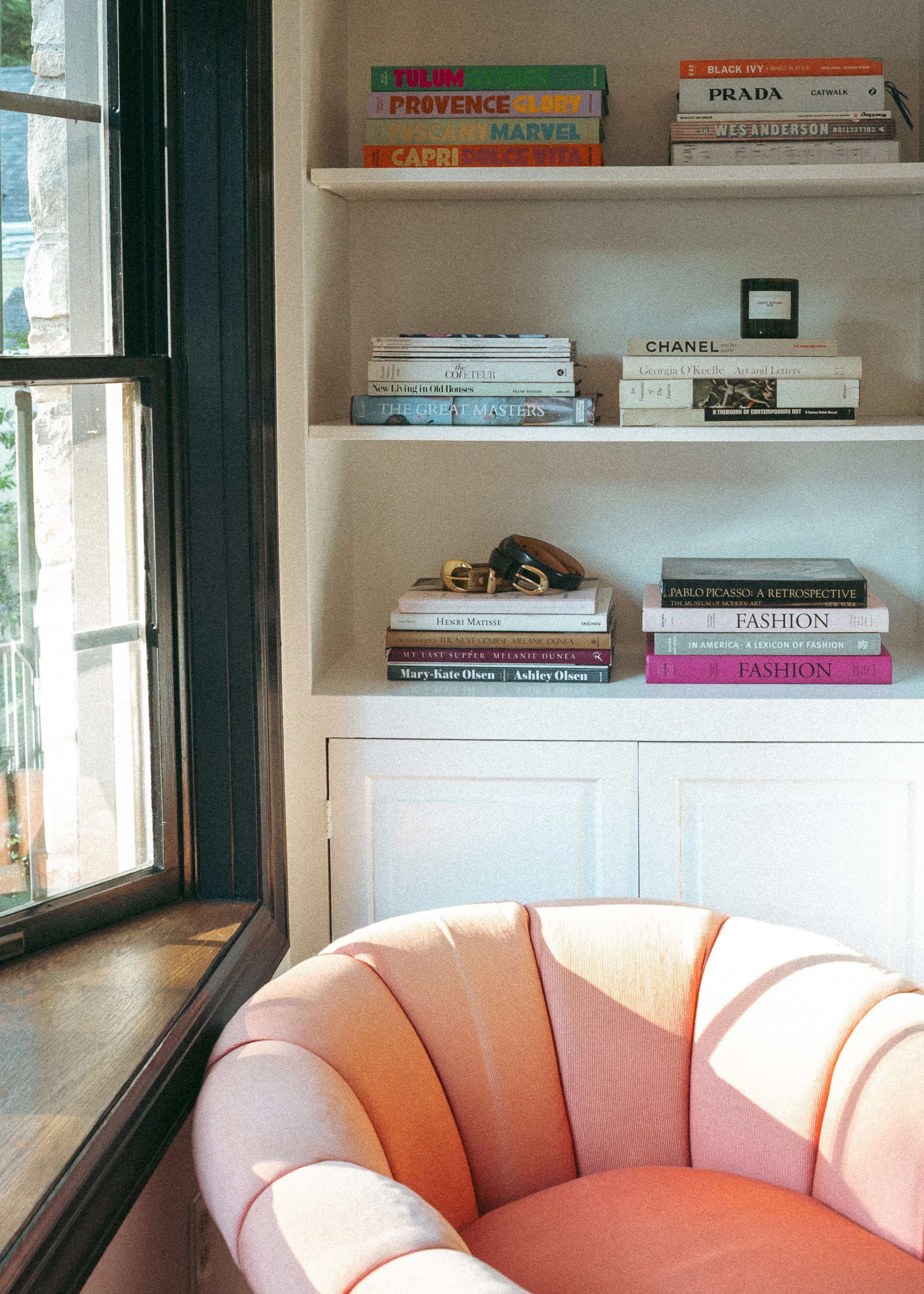 A bookshelf with art, fashion, and travel books, a black small object, and sunglasses on a white cabinet, next to a pink and orange upholstered chair by a window with sunlight.