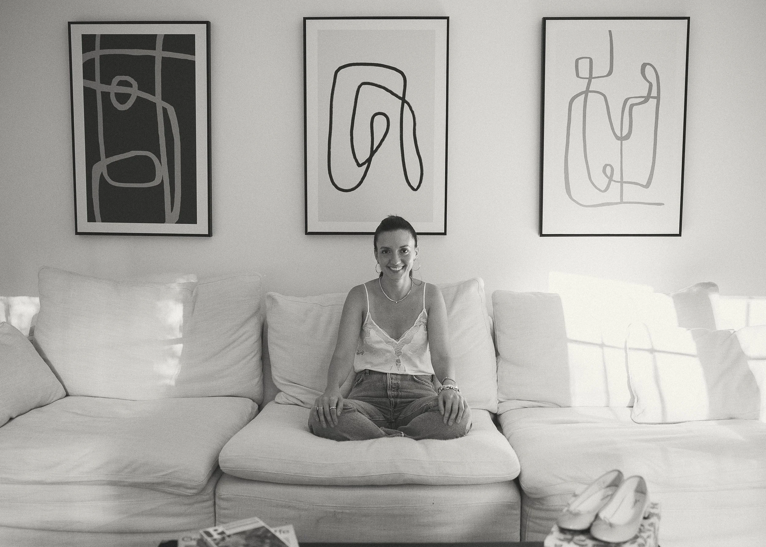 A woman with dark hair tied back, sitting cross-legged on a white sofa, smiling at the camera in a room with three abstract line art paintings on the wall behind her.