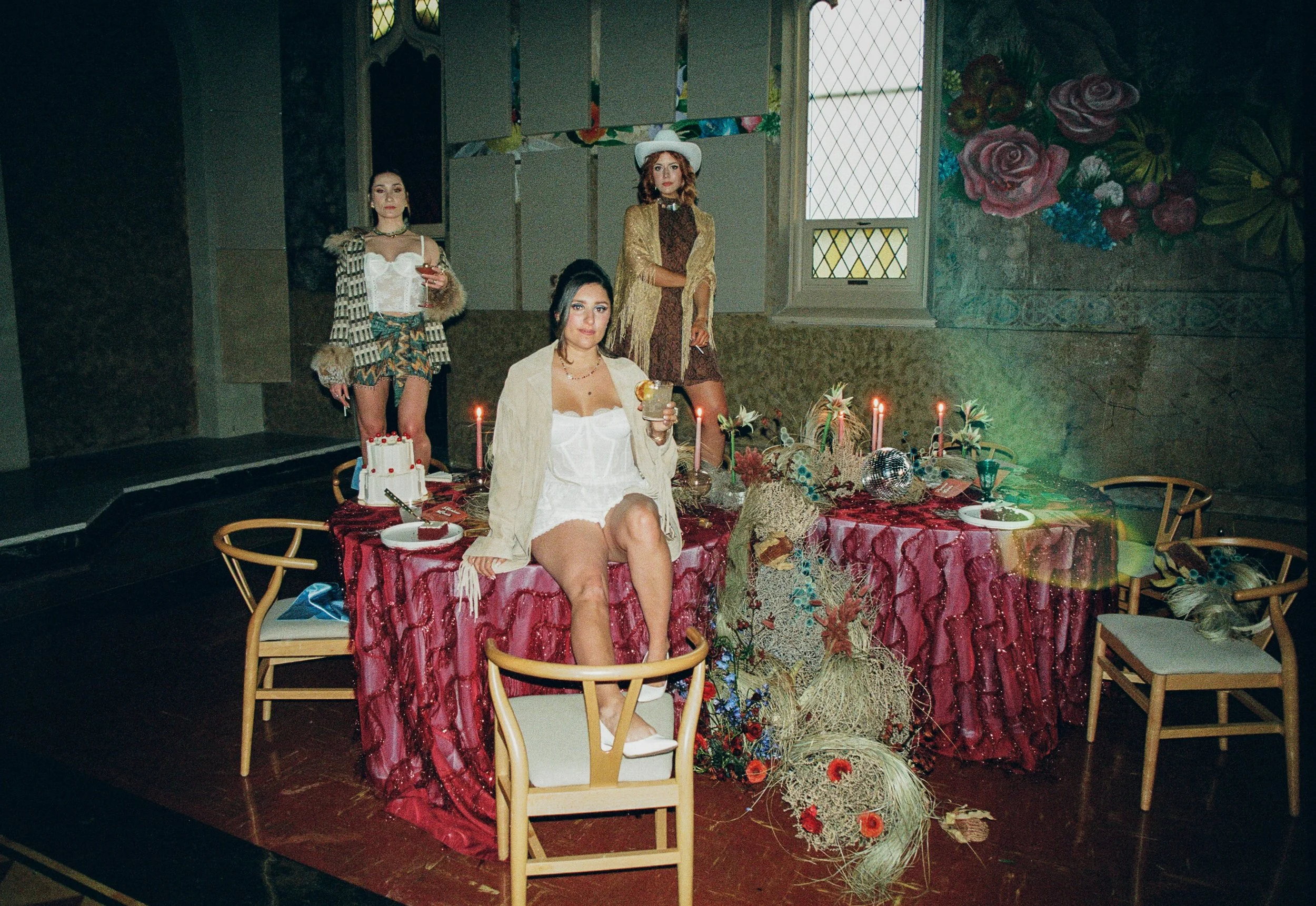 Four women at a decorated table with candles, cake, and flowers in a dimly lit room, with stained glass windows and floral wall art in the background.