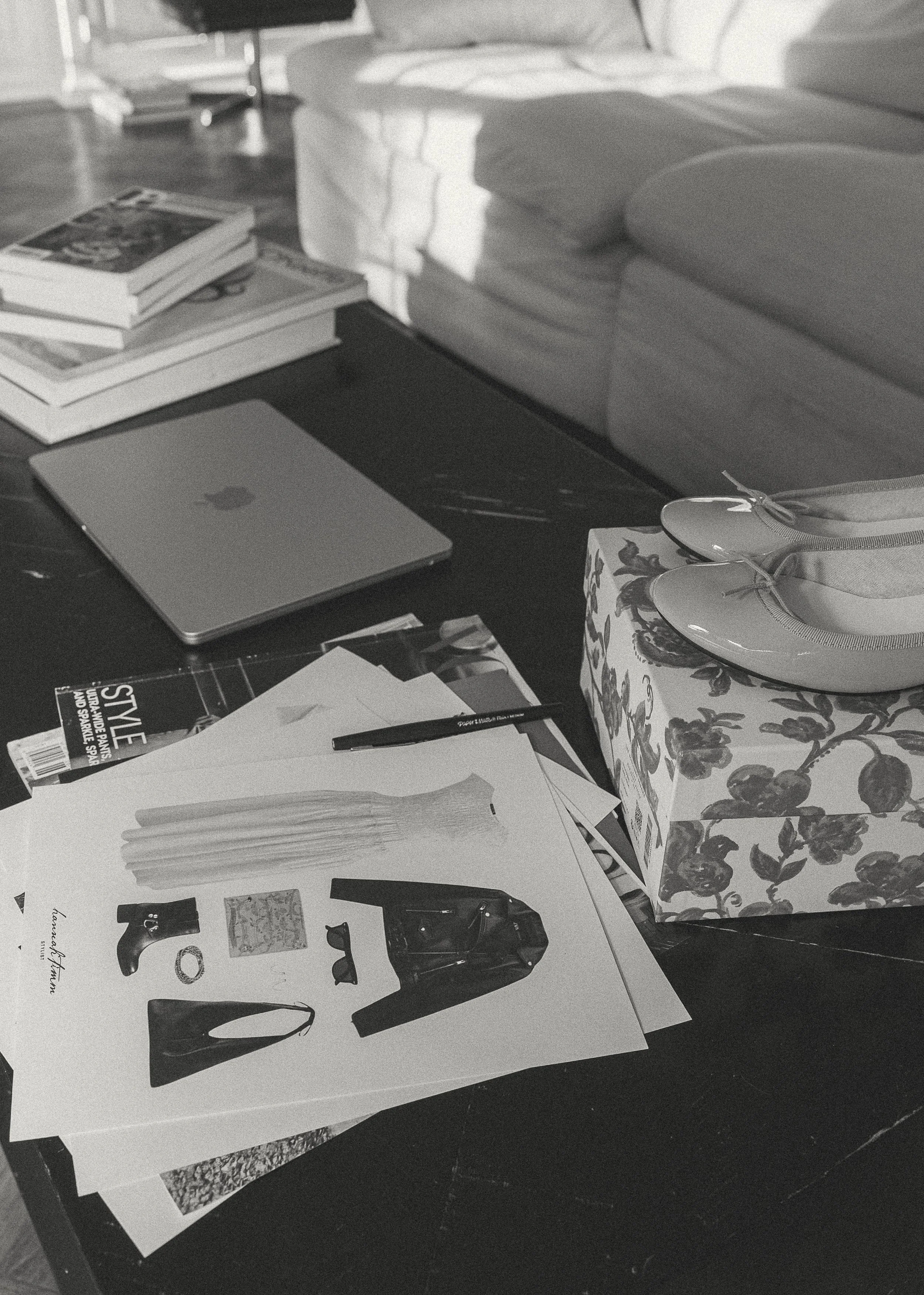 A cluttered coffee table with fashion design sketches, a closed laptop, stack of magazines, a pen, a box of shoes, and a magazine.