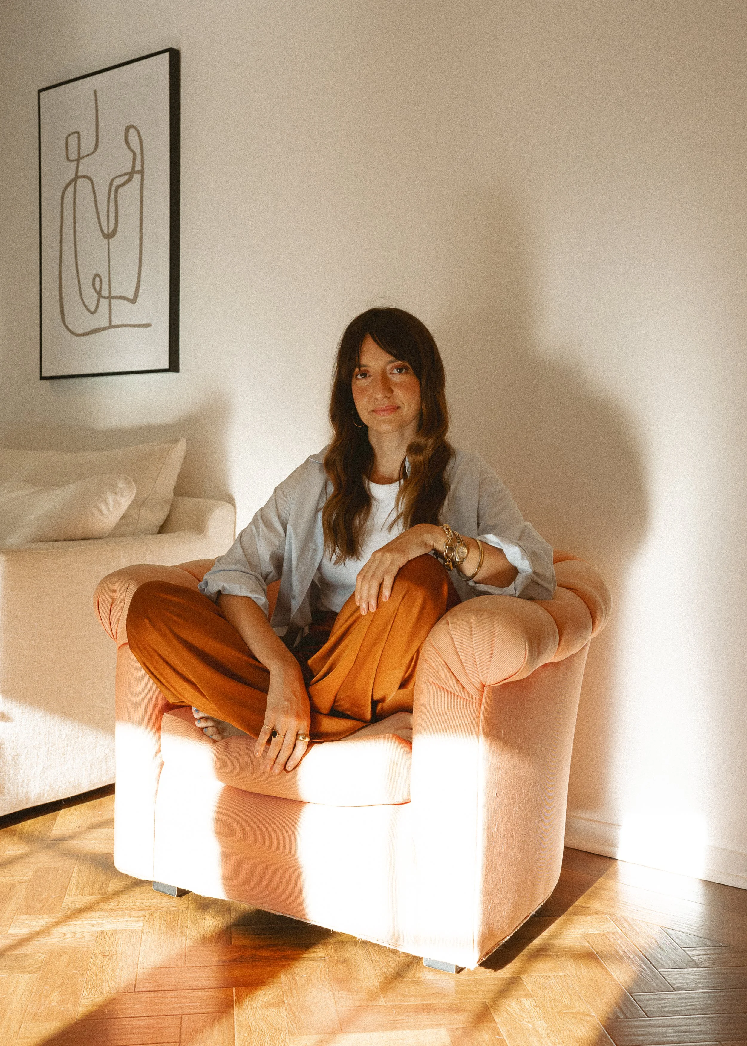 A woman with long wavy brown hair, sitting cross-legged on a peach-colored armchair in a room with white walls and wooden flooring. She is wearing a white top, which is partially covered by a light blue shirt, and orange-brown pants. She has accessories on her left wrist, a ring on her right hand, and is smiling slightly. There is a framed line art drawing hanging on the wall behind her, and part of a white sofa is visible on the left side of the image.