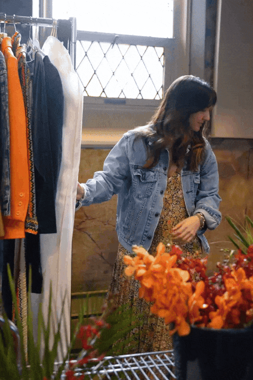 A woman shopping for clothes at a boutique, wearing a floral dress and denim jacket, with hanging clothes rack and a flower arrangement nearby.