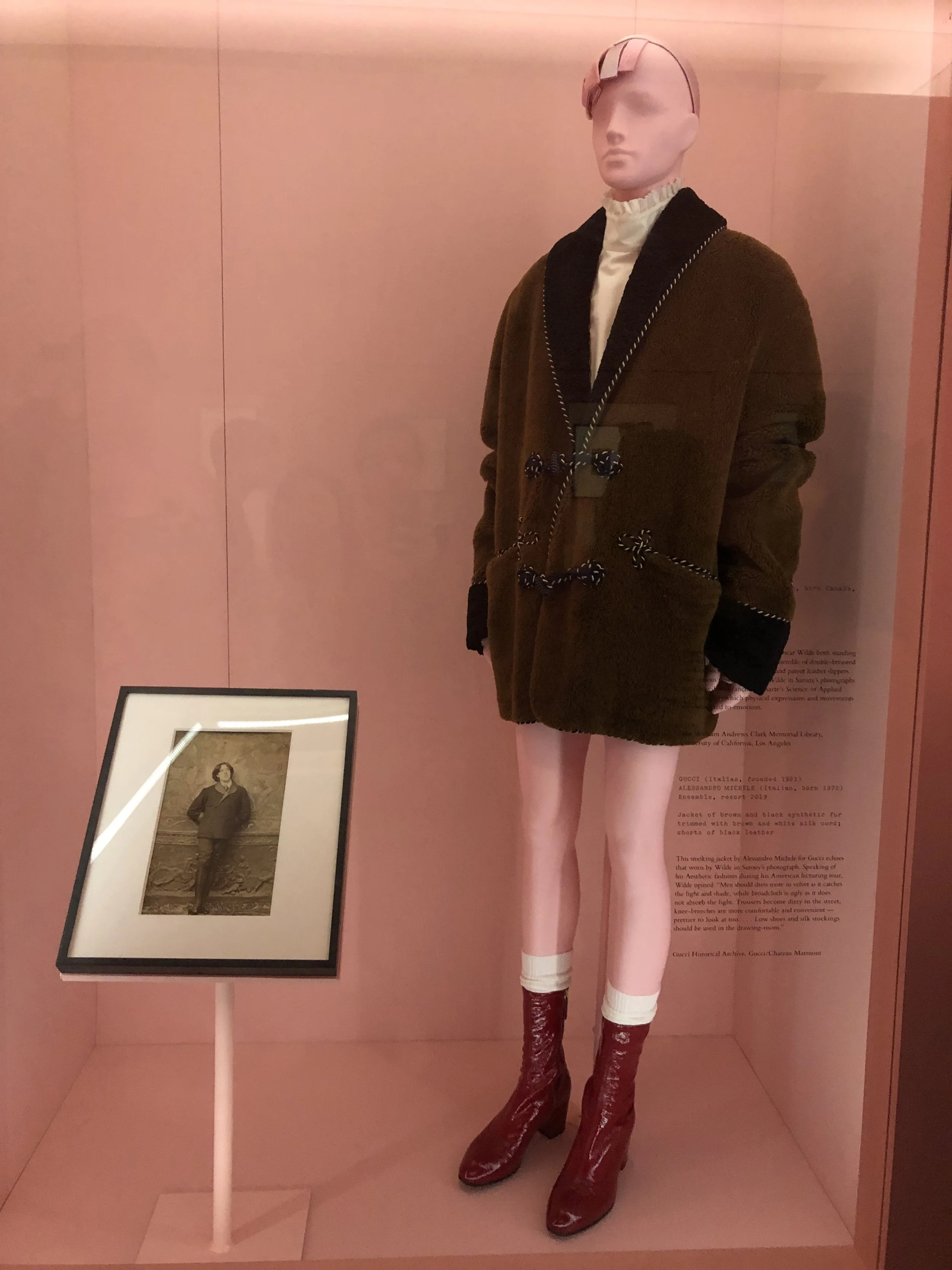 A mannequin dressed in a brown coat, white turtleneck, pink tights, and red shiny boots, displayed in a pink-colored exhibit with a photo and text on the background wall.
