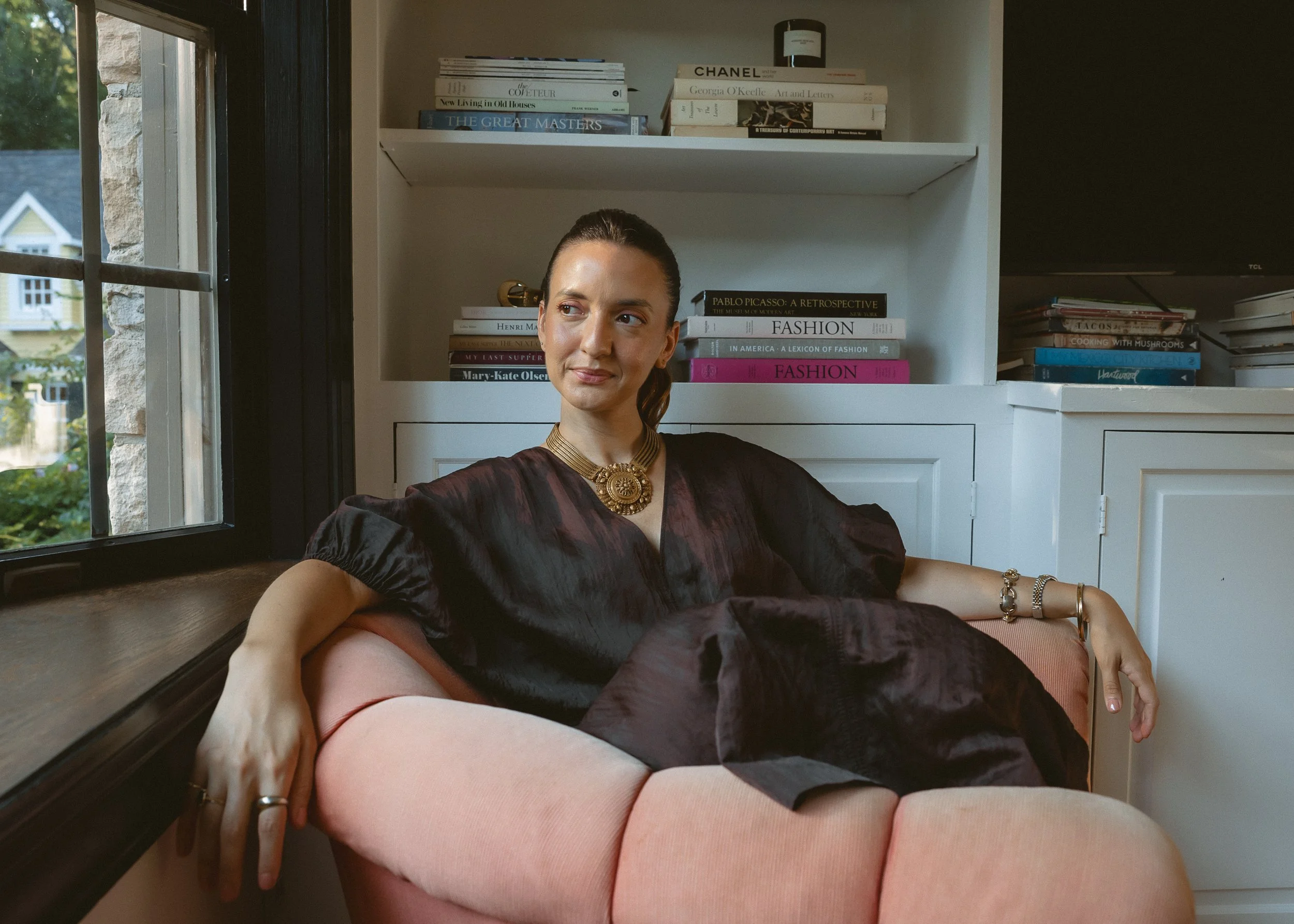 A woman sitting on a pink couch near a window, with a bookshelf behind her filled with books about fashion and art.