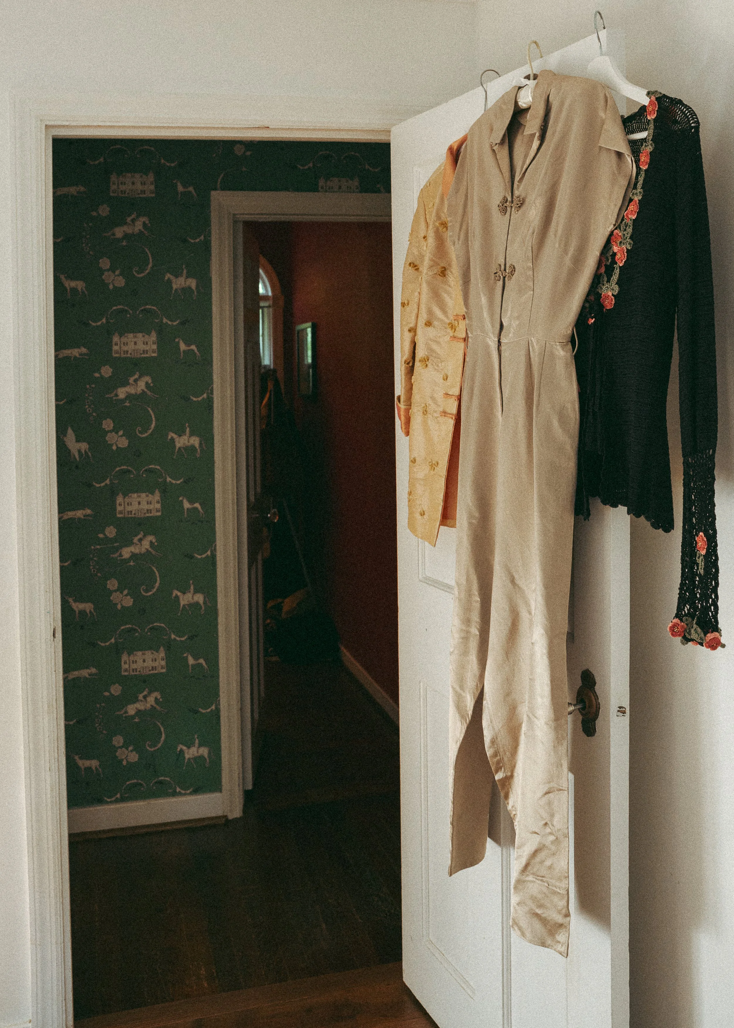 A closet door with a few dresses hanging on it, in a room with dark wood flooring and patterned wallpaper with horses and houses.