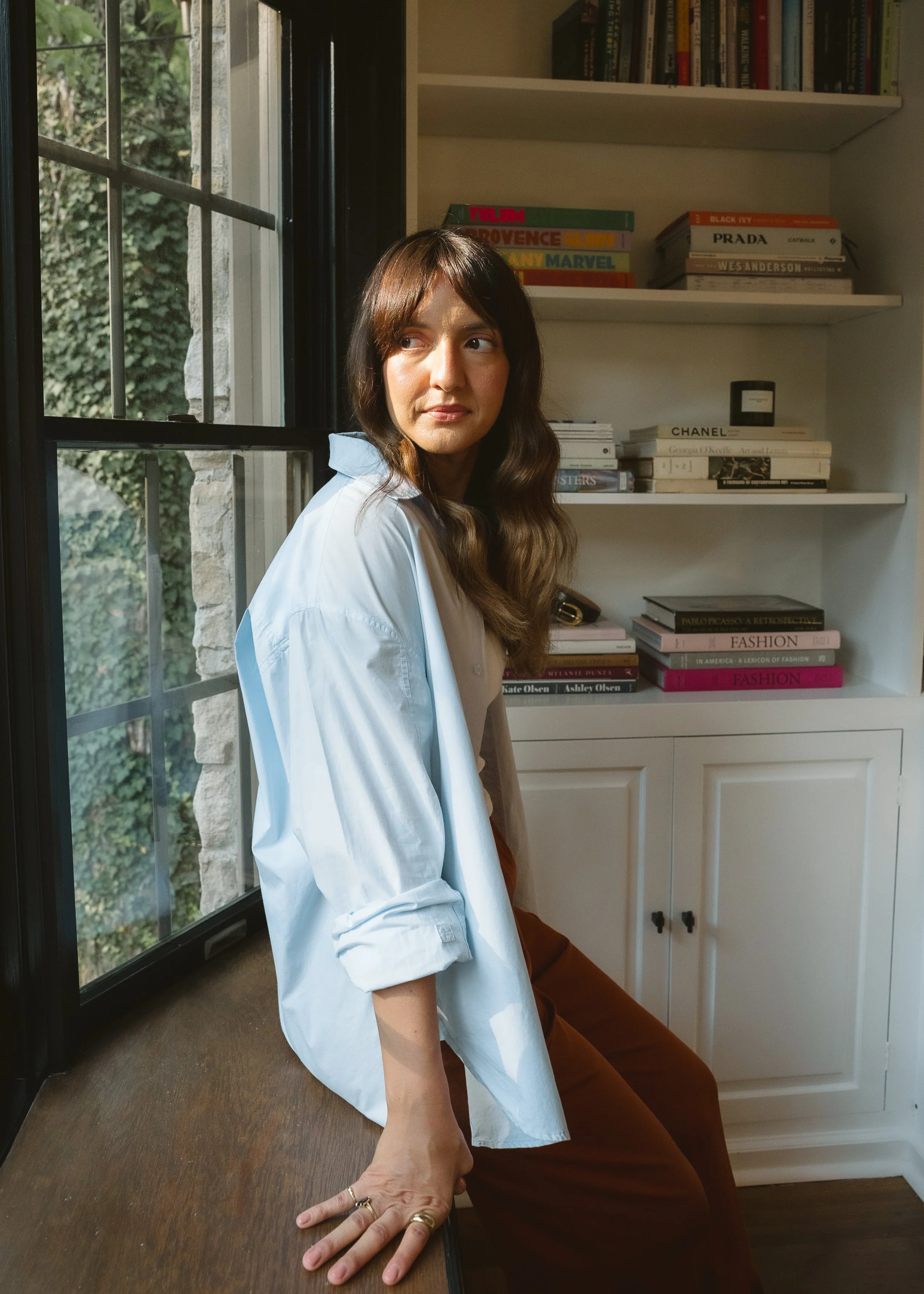 A woman with long brown hair and a white shirt sitting on a wooden surface next to a window, with a bookshelf filled with books behind her.