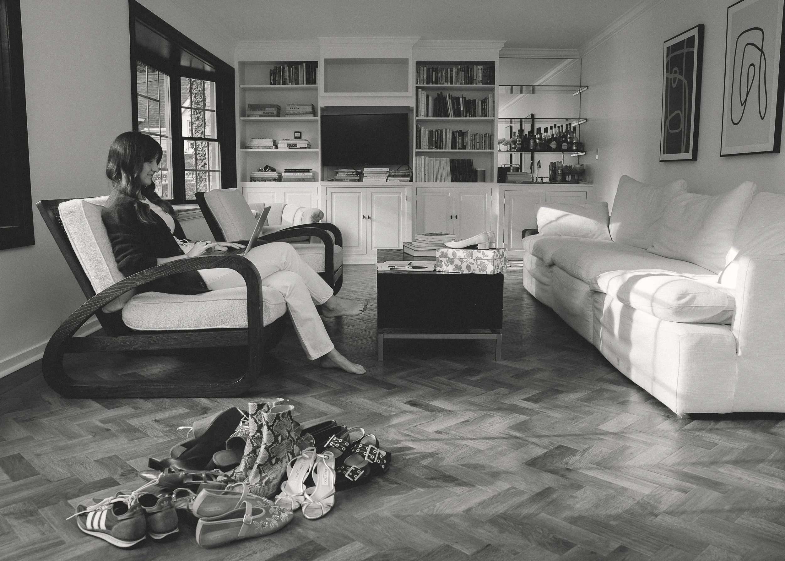 A woman sitting on a chair with a laptop in a living room with a white sofa, a coffee table, and a bookshelf on the wall. Shoes and bags are on the floor in front of her.