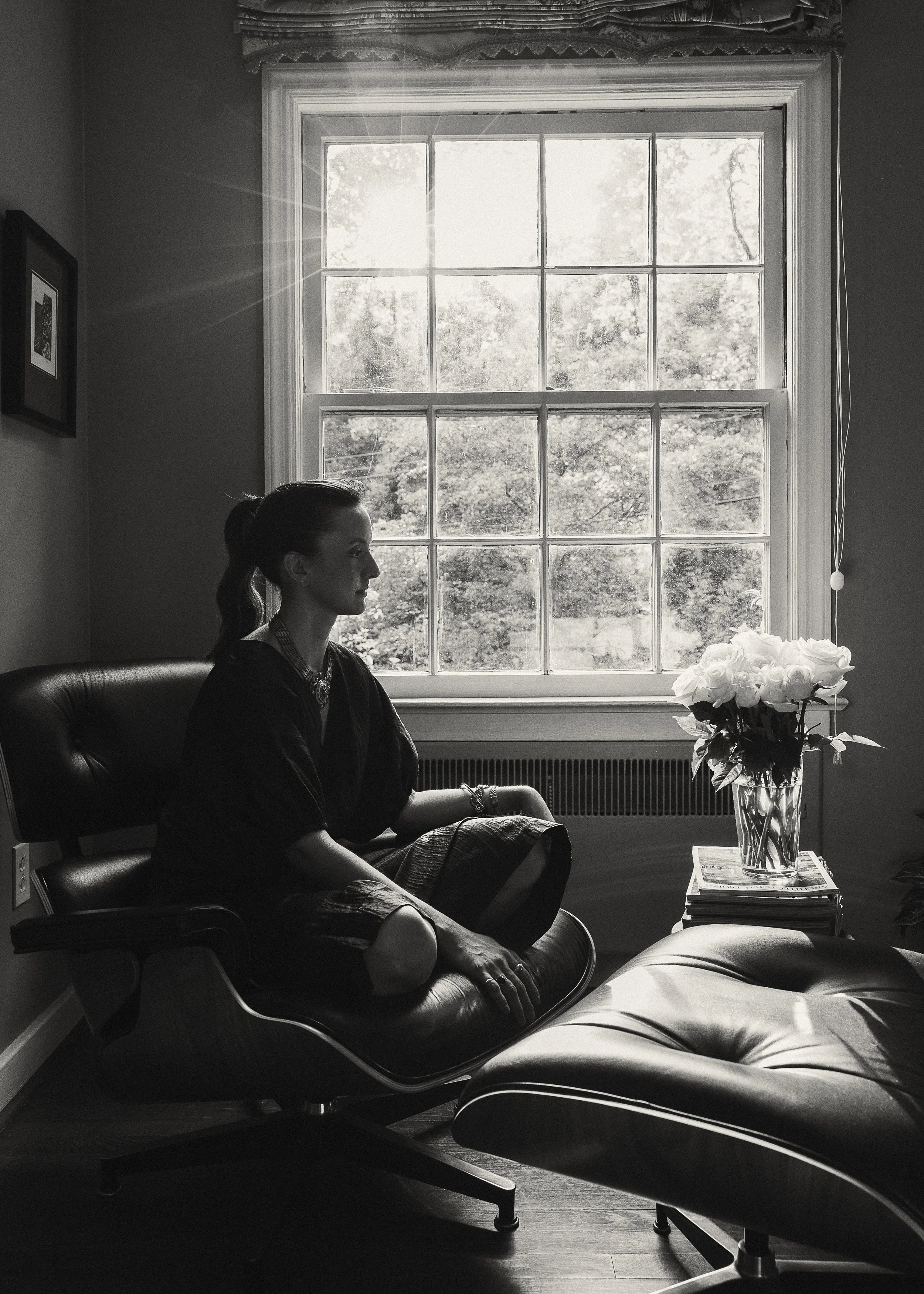 A woman sitting cross-legged on a leather lounge chair facing a large window, with a vase of flowers on a table beside her, in a black and white interior shot.