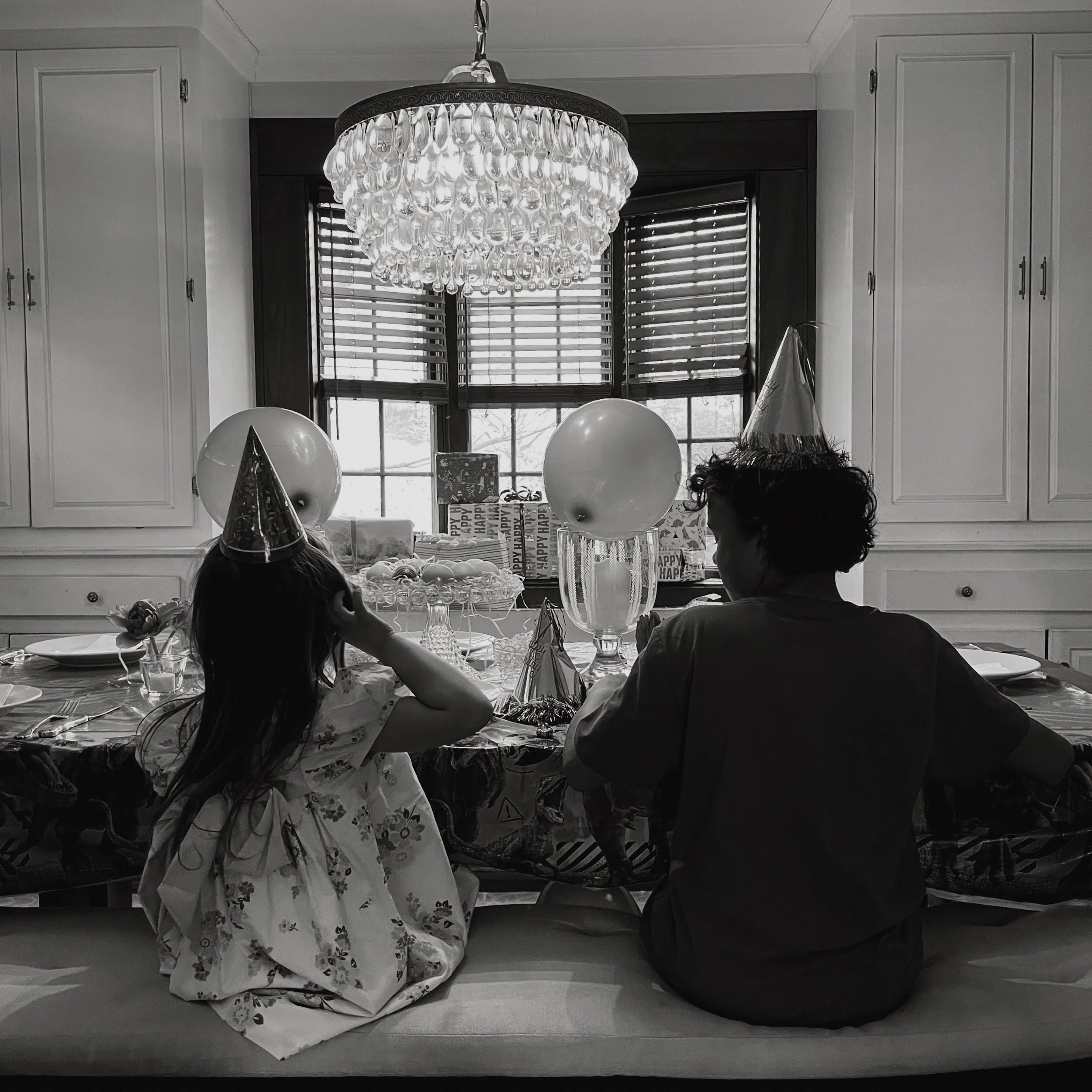 A child and an adult sitting at a decorated birthday table with balloons, party hats, and gifts in a dining room with a large window and a chandelier.