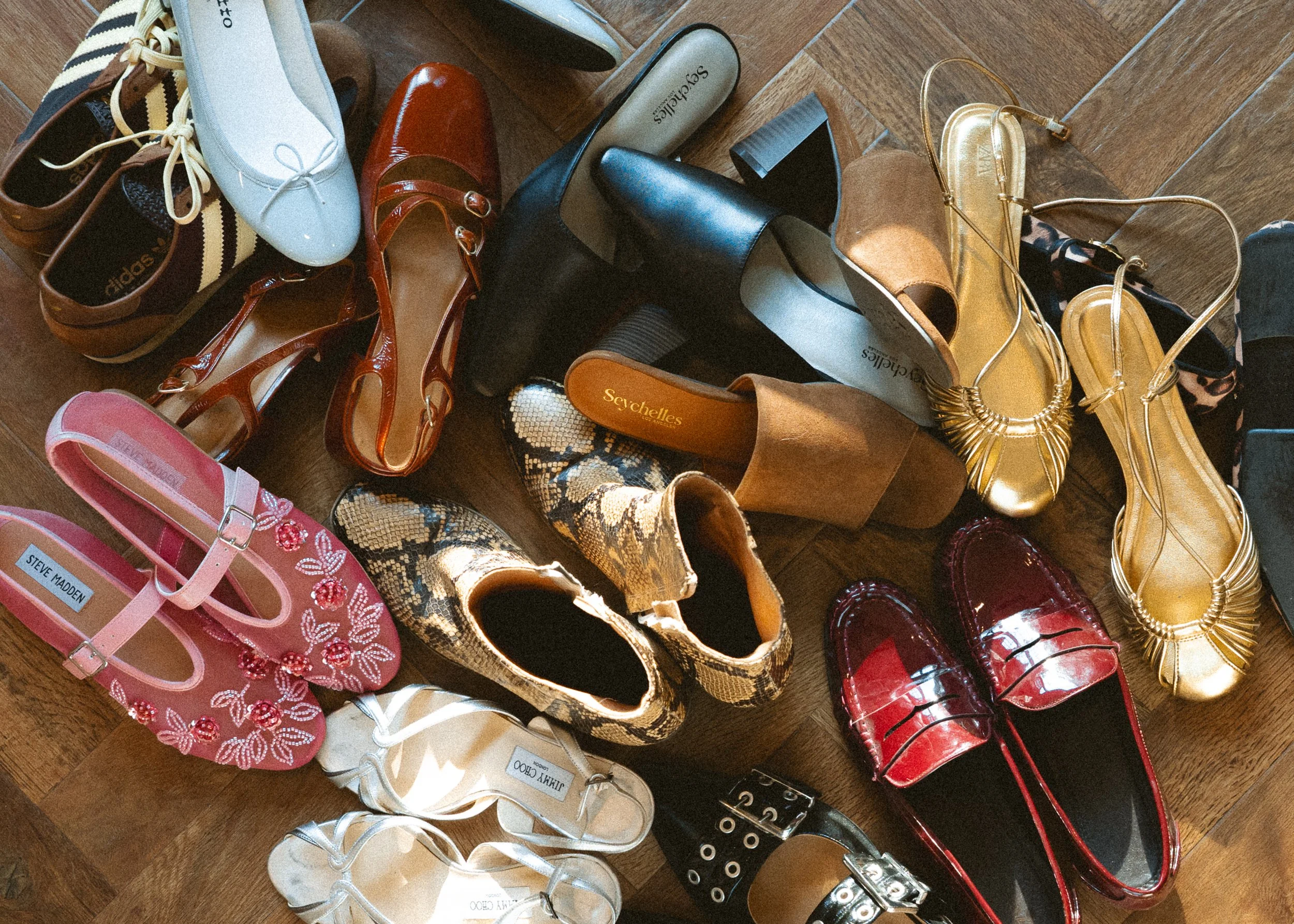 A variety of women's shoes arranged on a wooden floor, including sandals, high heels, flats, and boots in different colors and styles.