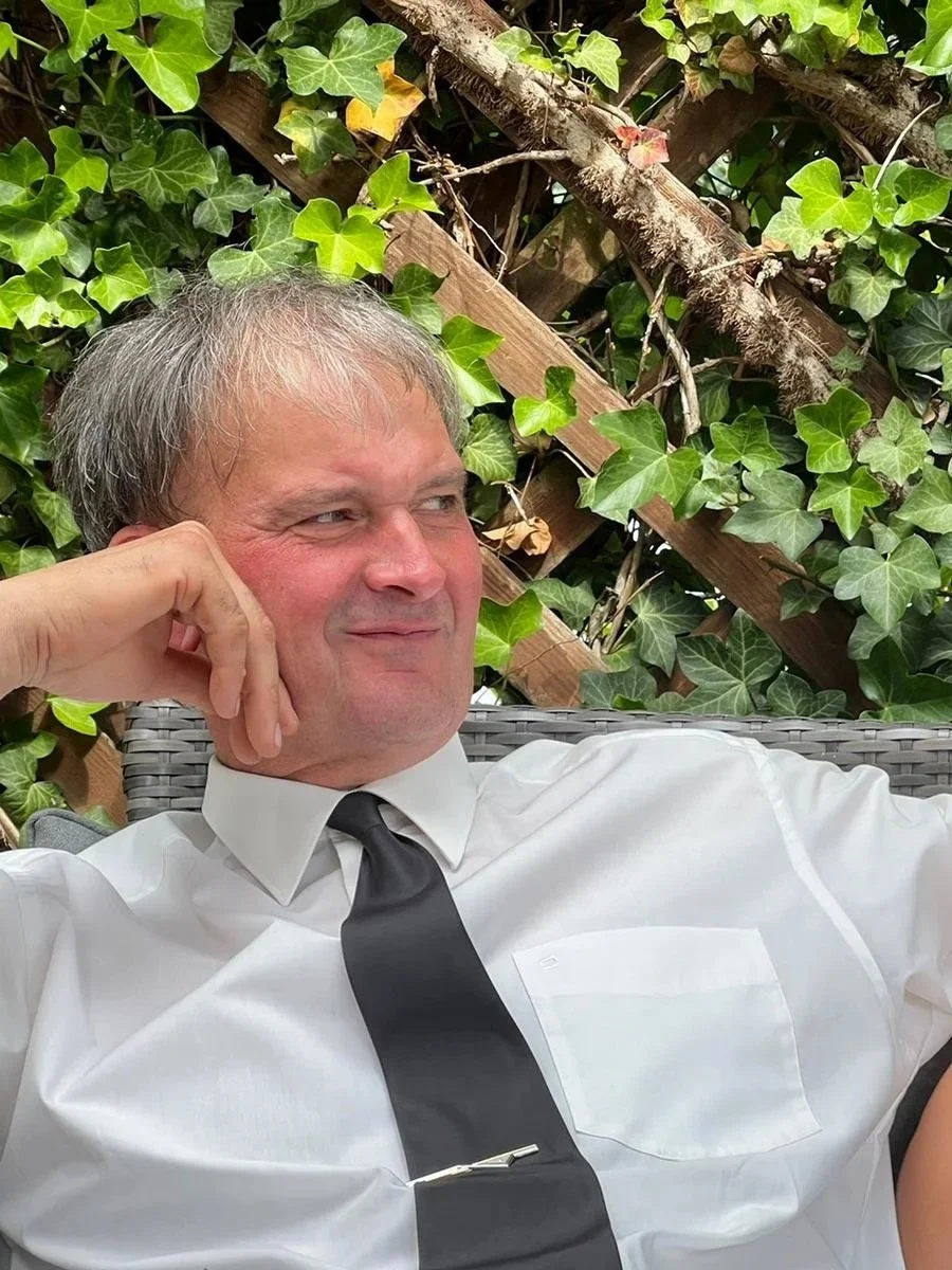 Mann in weißem Hemd und schwarzer Krawatte sitzt entspannt vor einer Kletterpflanze mit großen grünen Blättern.