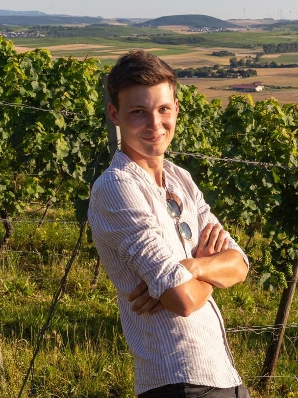 Junger Mann steht in einem Weinberg mit grünen Reben, im Hintergrund eine ländliche Landschaft mit Feldern und Hügeln bei Sonnenuntergang.