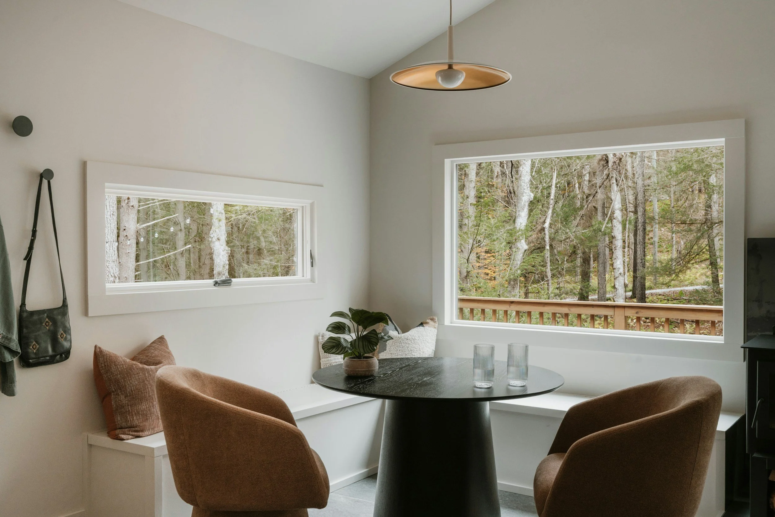 Portland staged Interior of a cozy dining nook with a round black table, two brown upholstered chairs, and a large window showing a wooded outdoor scene. There's a side bench with cushions, a plant on the table, and a ceiling light fixture.
