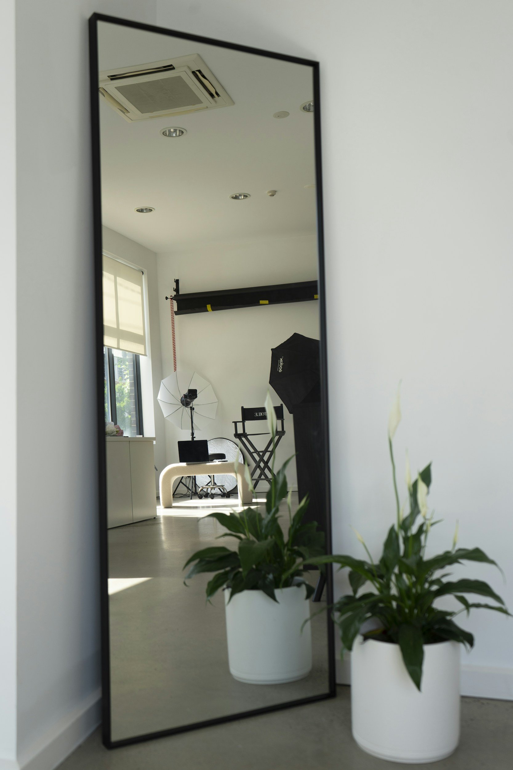 Reflection of a photography studio setup in a full-length mirror, with lights, equipment, a chair, and potted plants nearby.