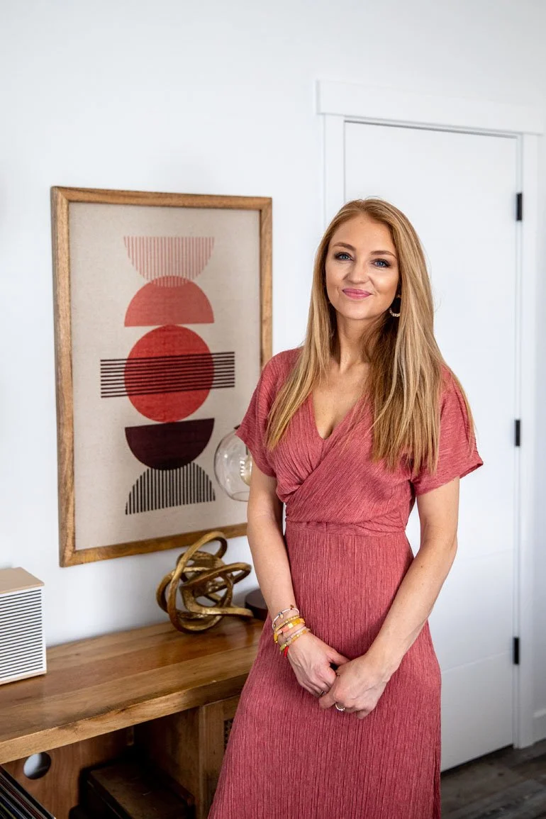A woman with long blonde hair in a pink dress standing indoors next to a wooden table, with abstract wall art and decorative objects in the background.