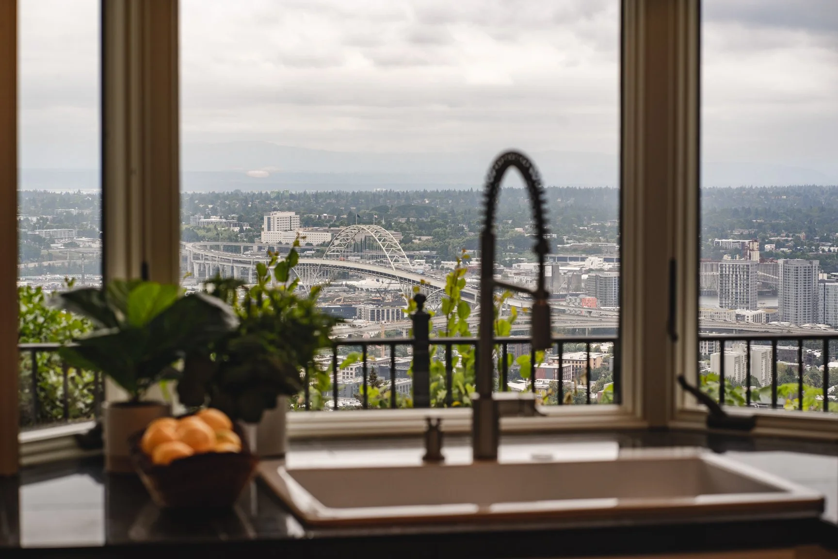 Living room with a tan leather sectional sofa, patterned throw pillows, a black coffee table with books and a plant, a black open shelving unit with books and decorations, a large potted plant, a textured rug, and a mirror on the wall reflecting a window.