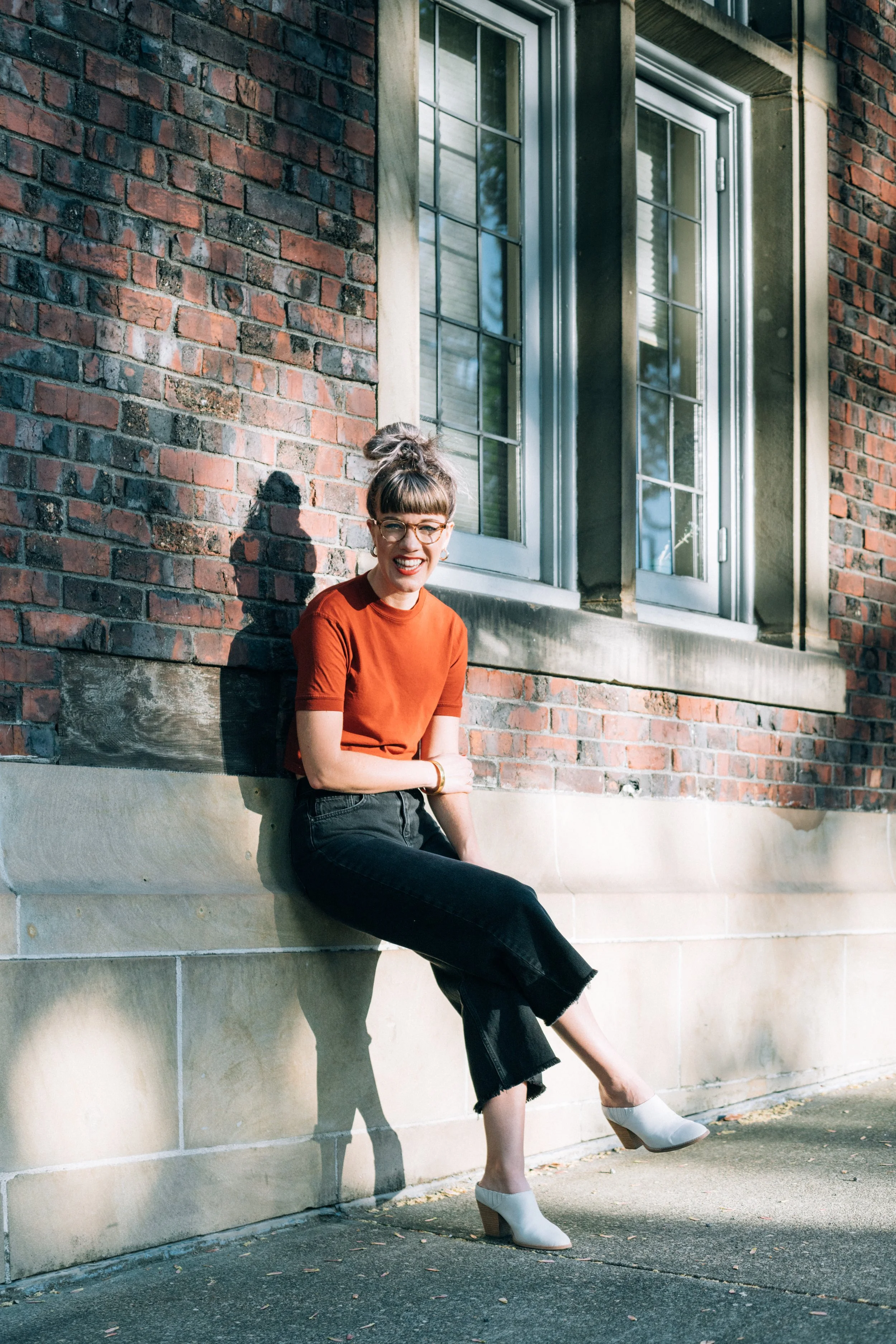 A woman sitting on a stone ledge outside of a brick building, smiling. She is wearing a red top, black jeans, and white heeled shoes. Sunlight casts a shadow on the brick wall.
