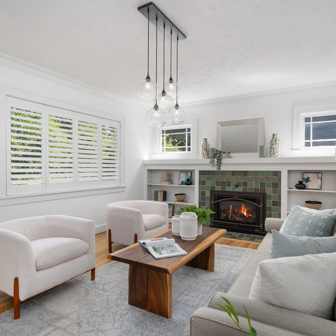 Living room with a brown leather sofa, decorative pillows, and a white blanket. A round wooden coffee table with a sculpture and a book. A yellow ottoman, a side table with a black lamp, a bouquet of flowers, and a large leafy plant. The wall behind has multiple framed art pieces, including desert landscape photos, line art, abstract designs, and a black-and-white photo.