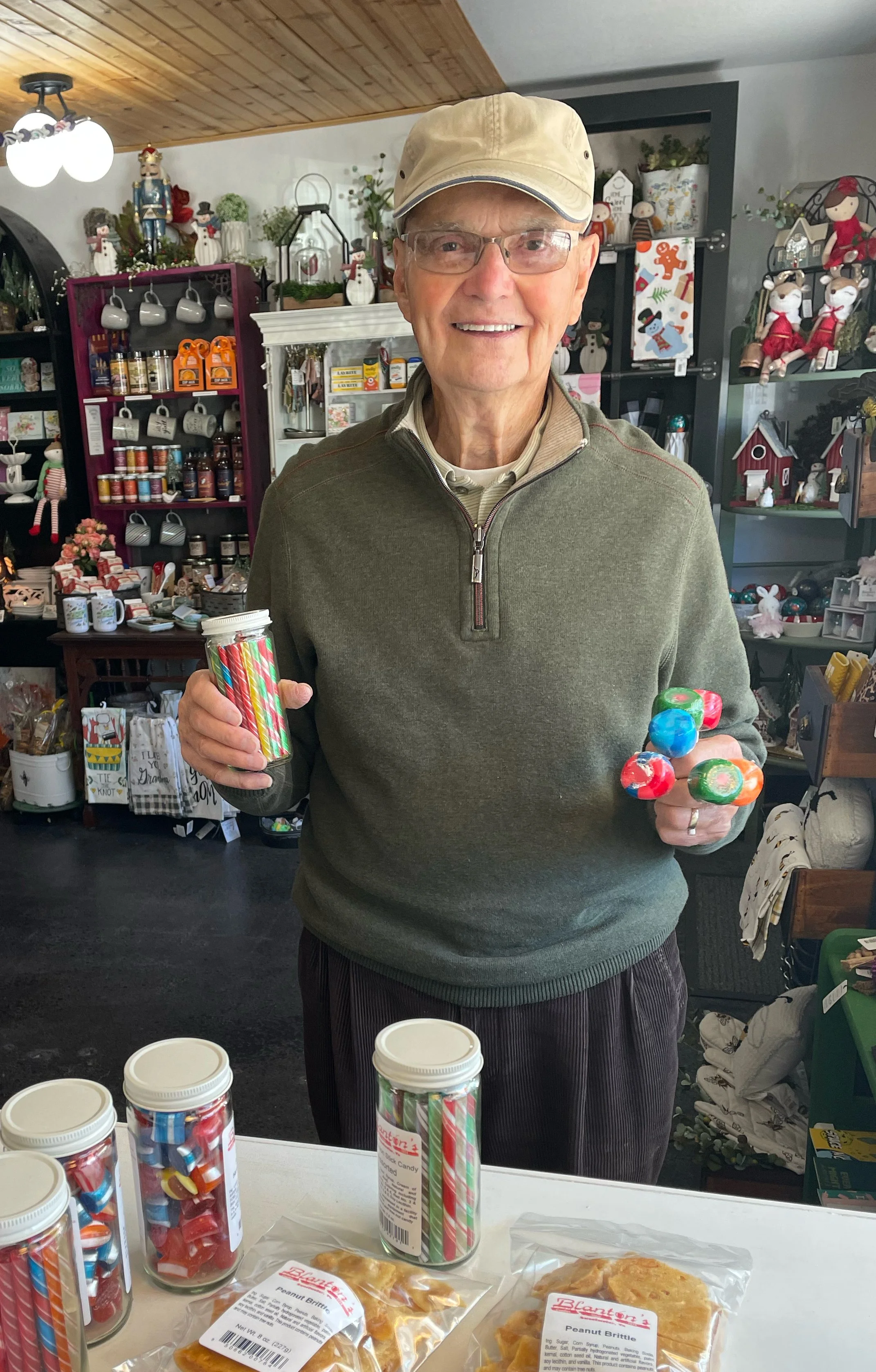 An elderly man wearing glasses, a beige cap, and a green quarter-zip sweater stands behind a table with holiday treats and candy jars. He is smiling and holding a handful of colorful round candies in one hand and a container of striped holiday candies in the other. The background is decorated with Christmas-themed ornaments and decorations.