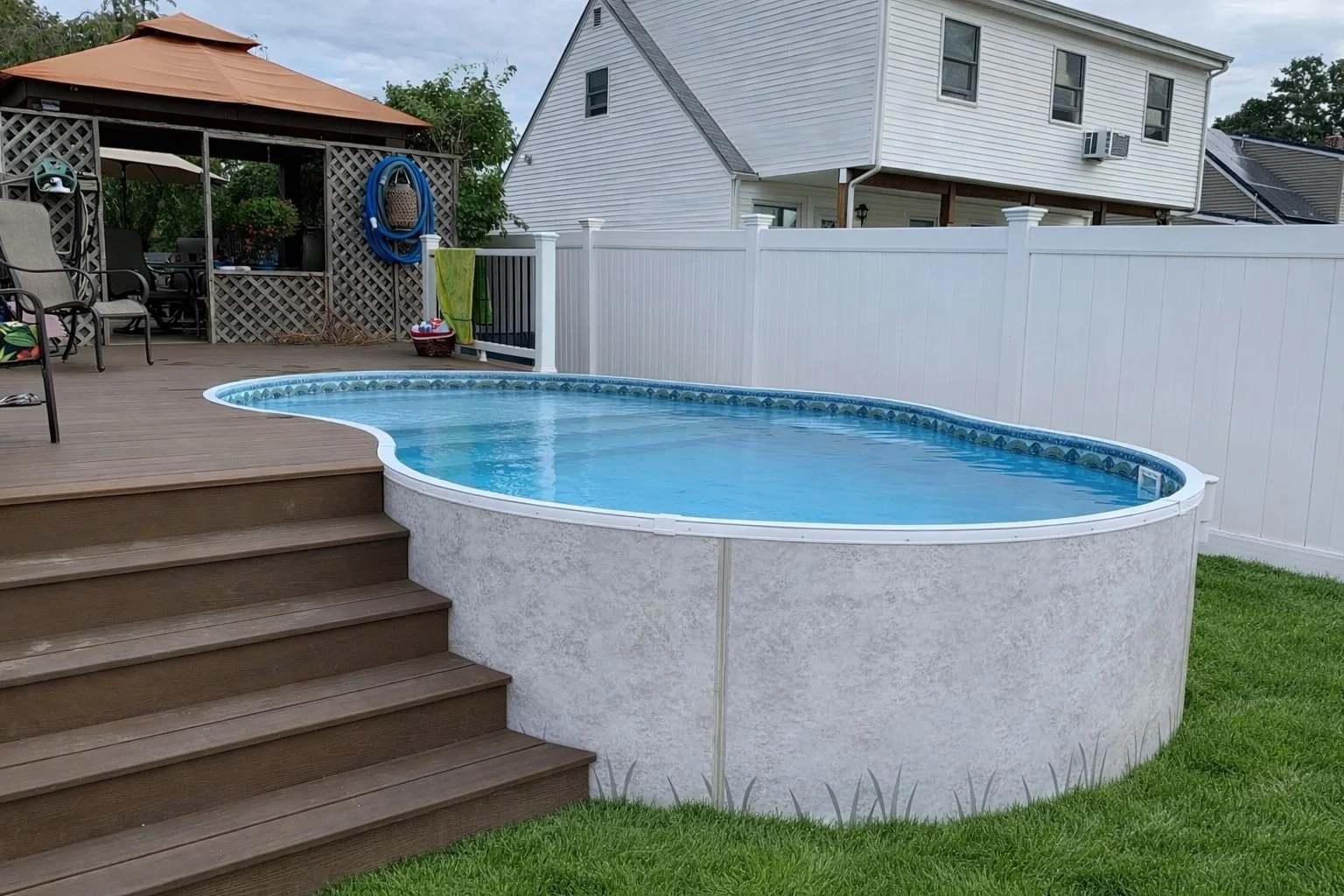 Backyard with above-ground swimming pool with wooden steps, surrounded by a white fence, and outdoor furniture including chairs and a patio table under a gazebo.