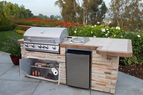 Outdoor outdoor kitchen with a stainless steel grill, mini fridge, and stone countertop, set against a garden with colorful flowers and trees in the background.