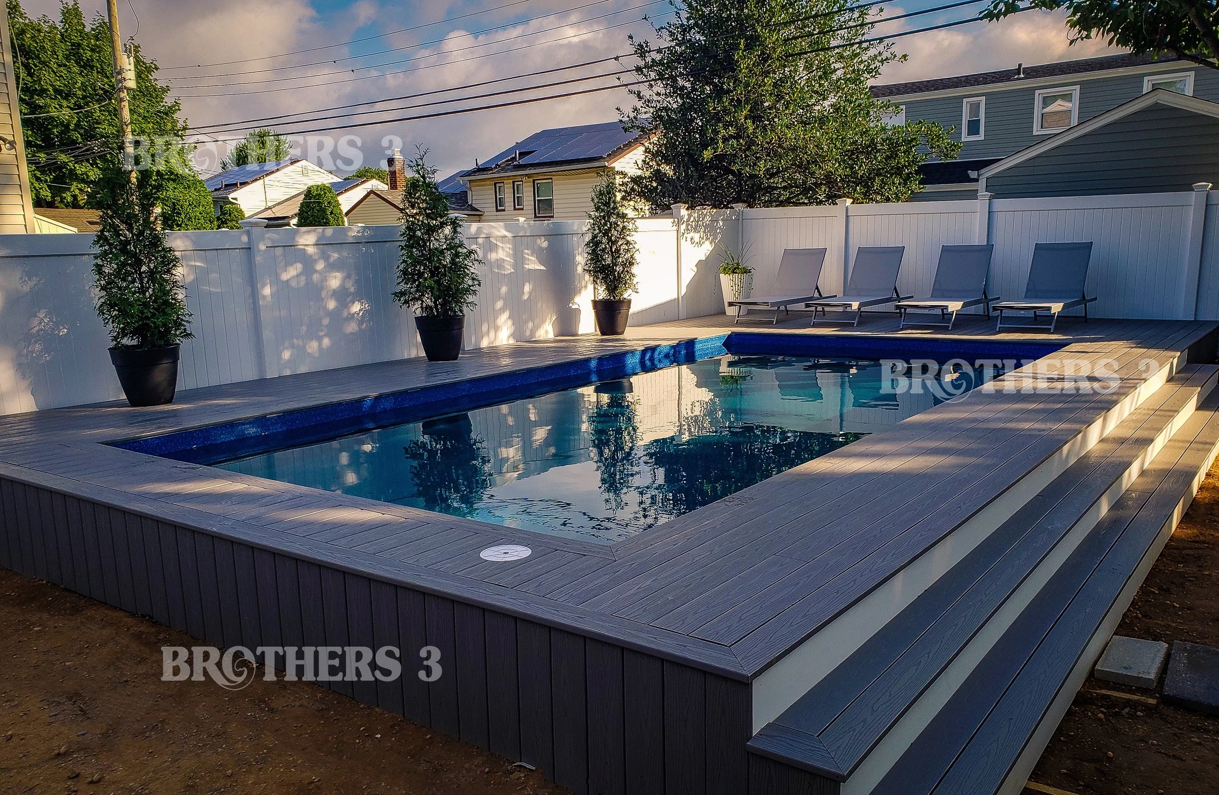 Residential backyard with a rectangular swimming pool surrounded by a wooden deck, white privacy fence, potted plants, and four lounge chairs. Houses and power lines are visible in the background, and the sky is partly cloudy with sunlight reflecting on the water.