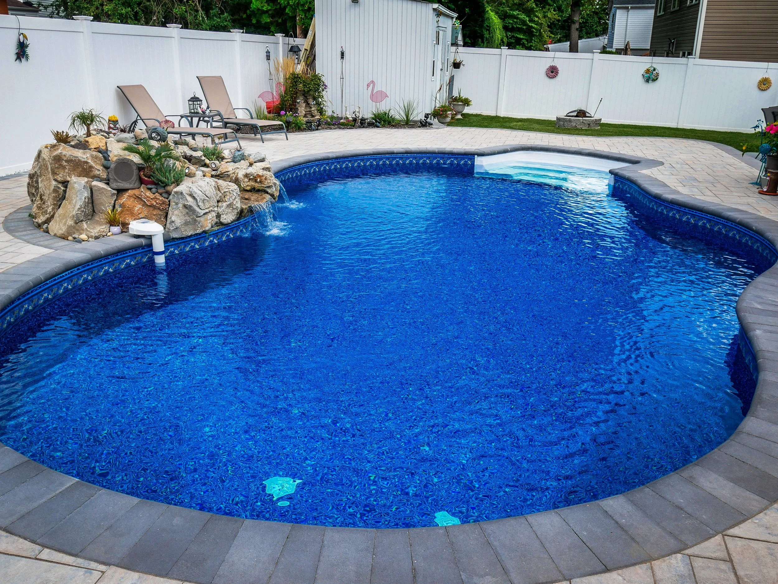 In-ground backyard swimming pool with blue water, surrounded by paving and a white fence-lined yard with decorated garden accents, chairs, and a small artificial waterfall feature.