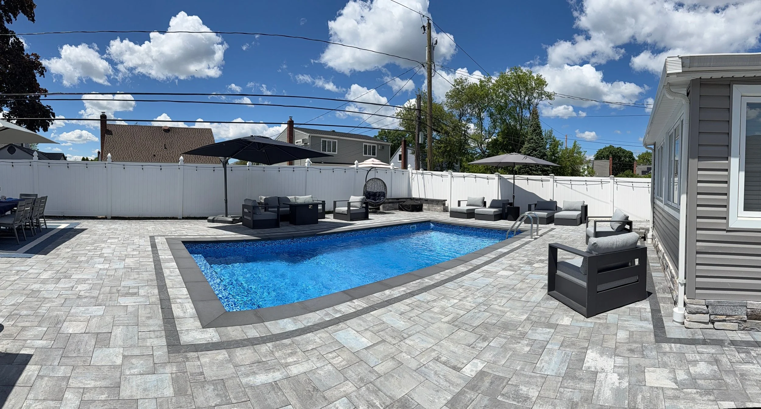 Backyard with a swimming pool, patio furniture with umbrellas, in-ground blue pool, white vinyl fence, house on the right, and a clear blue sky with scattered clouds.
