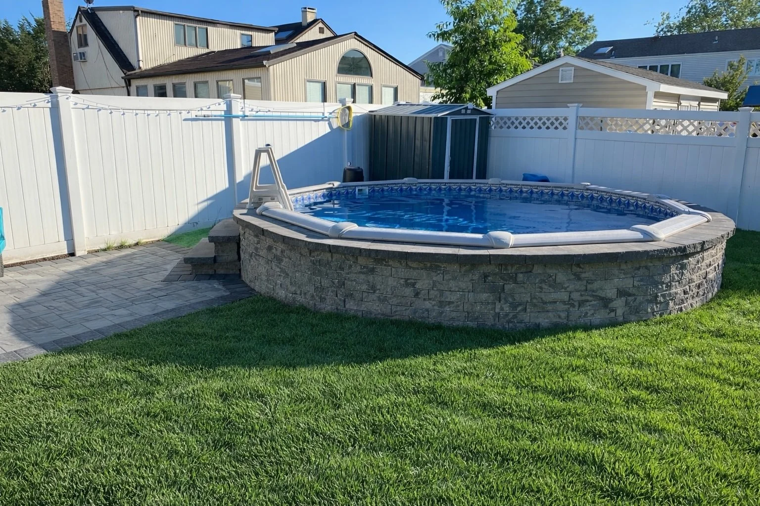 Backyard with above-ground swimming pool, white privacy fence, green grass, paved patio, and neighboring houses.